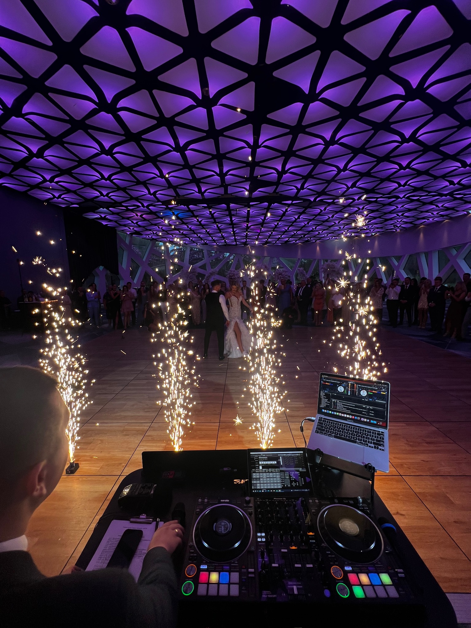 Para młoda tańczy na parkiecie, otoczona fontannami iskier i gośćmi. W tle widoczny sprzęt DJ-a, a nad głowami dekoracyjne oświetlenie w kolorze fioletowym.