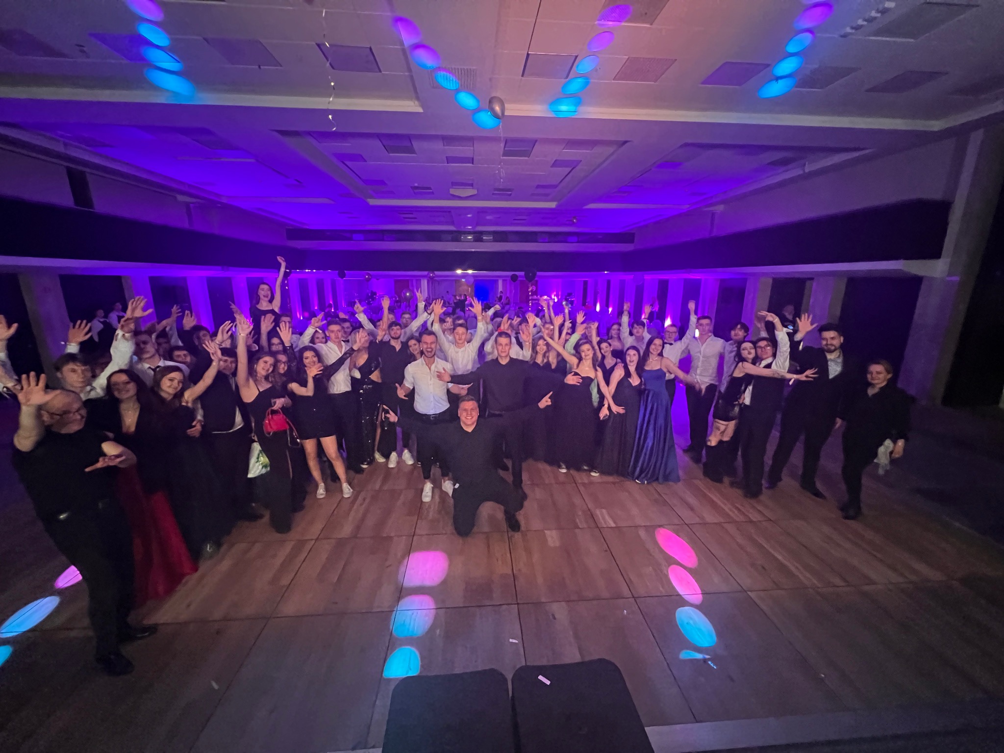Uczestnicy imprezy z podniesionymi rękami, oświetleni kolorowymi światłami na parkiecie. Osoba w czarnym stroju klęczy na pierwszym planie.