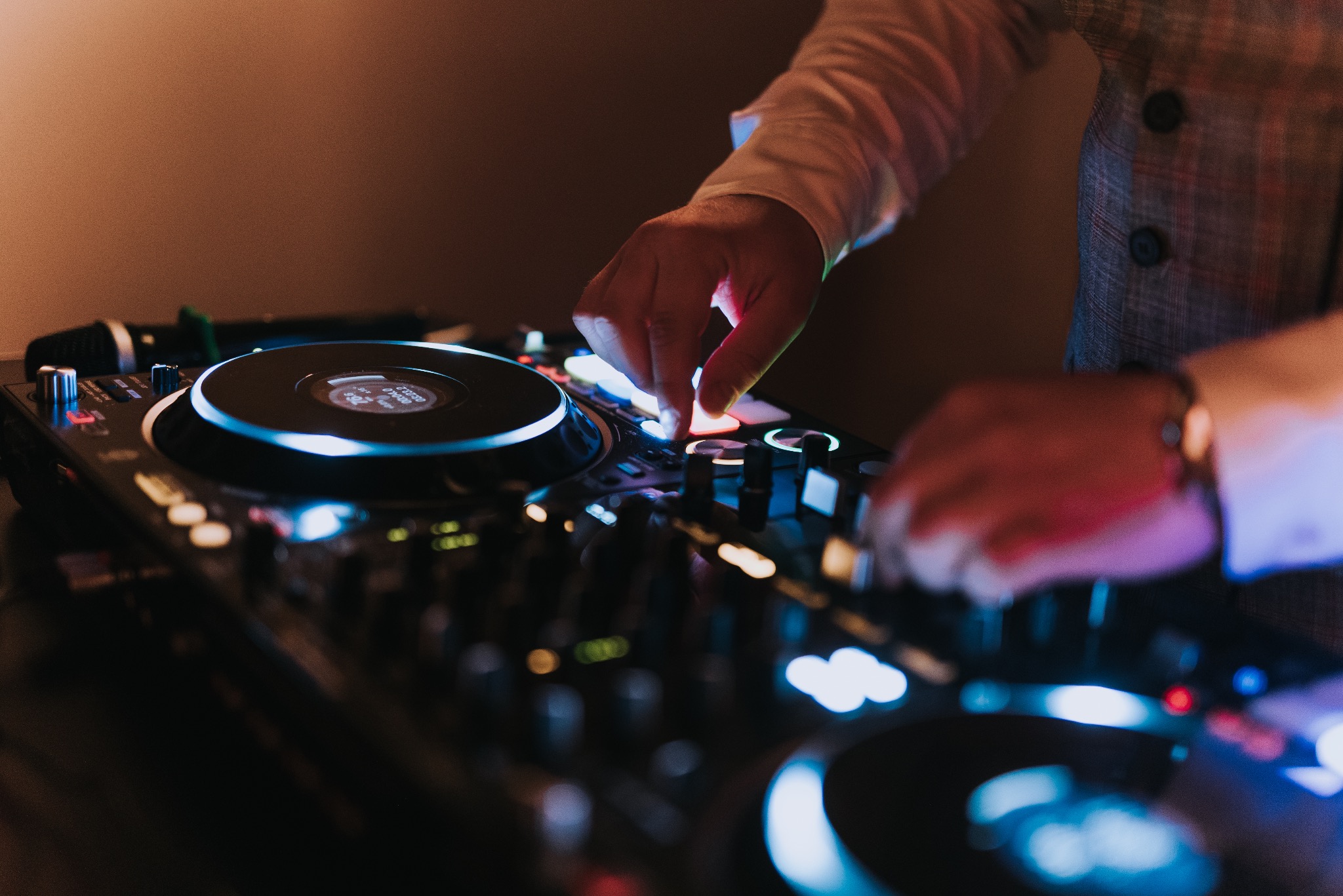 DJ w trakcie występu, miksujący muzykę na profesjonalnym sprzęcie. Widoczne dłonie na kontrolerze, mikrofon w tle i oświetlenie sceniczne.