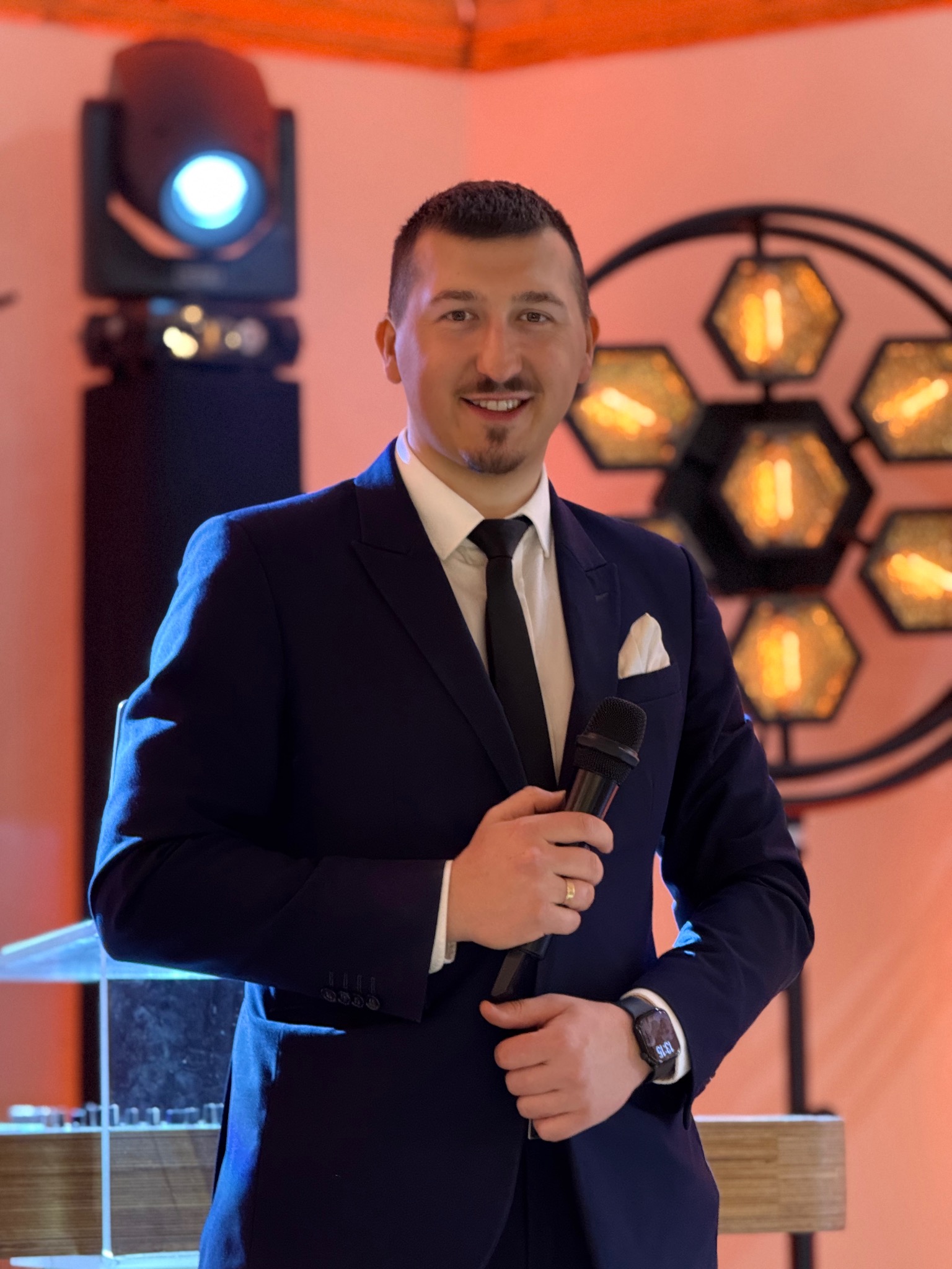 Uśmiechnięty konferansjer w granatowym garniturze trzyma mikrofon na tle oświetlenia scenicznego i dekoracji z żarówkami w kształcie plastra miodu.