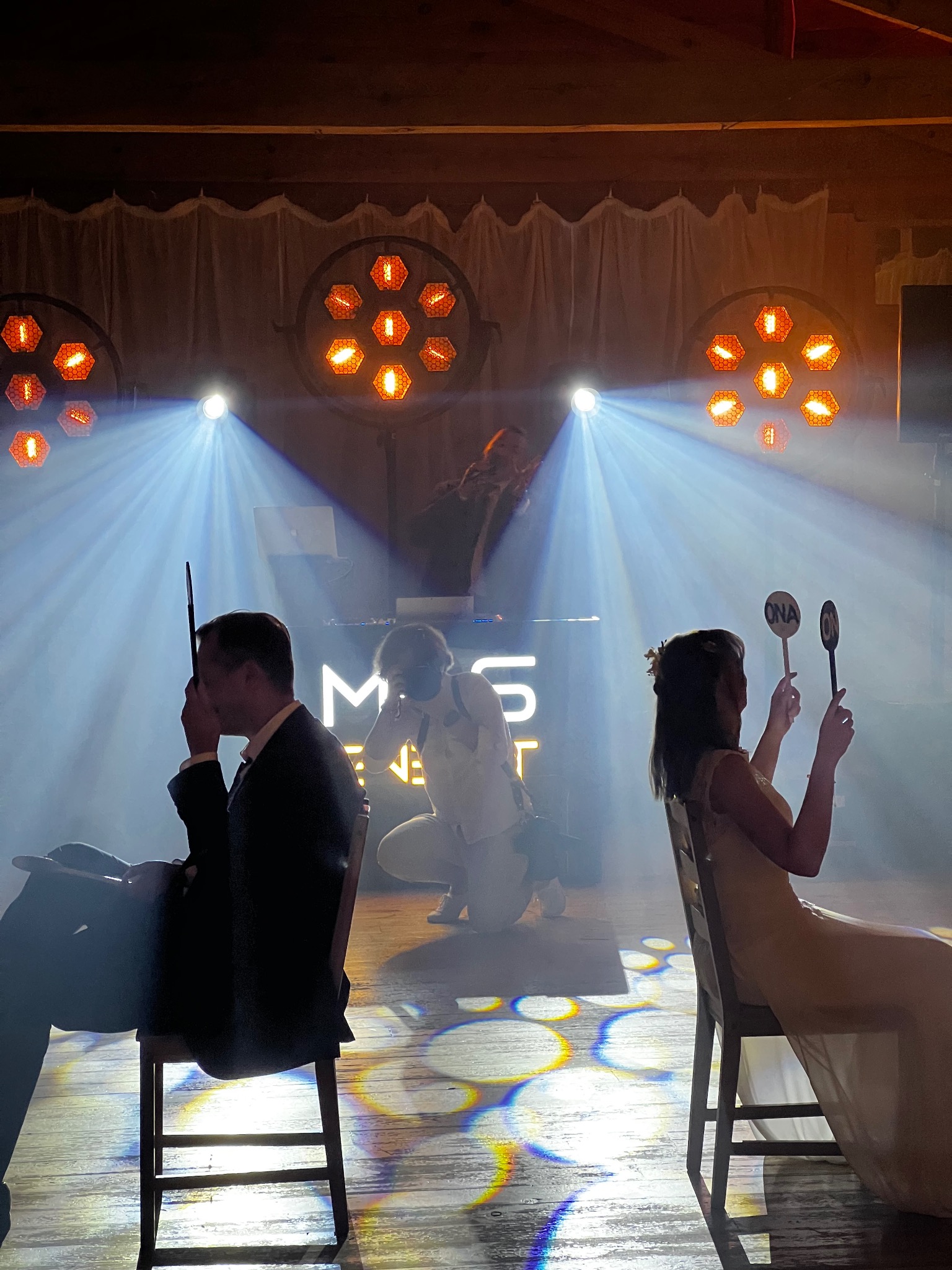 Para siedzi na krzesłach z rekwizytami 'ON' i 'ONA' w dłoniach, w tle DJ, fotograf i oświetlenie sceniczne, tworząc unikalną atmosferę wydarzenia.