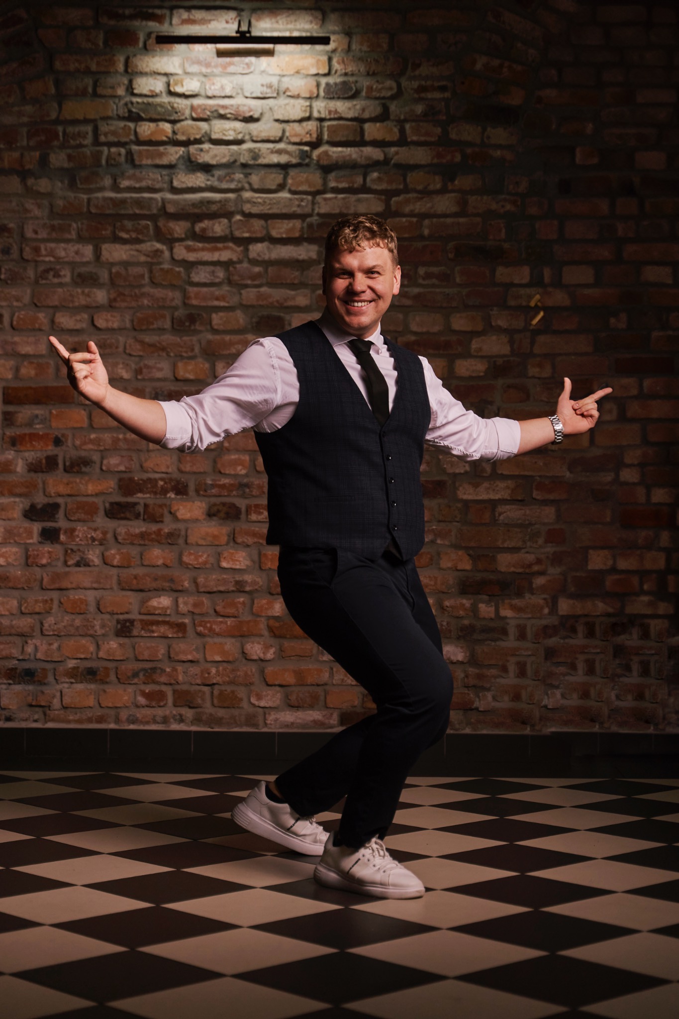 Uśmiechnięty mężczyzna w tańcu, w kamizelce i białych butach, na tle ceglanej ściany i szachownicy na podłodze. Dynamiczna poza, gestykulacja.