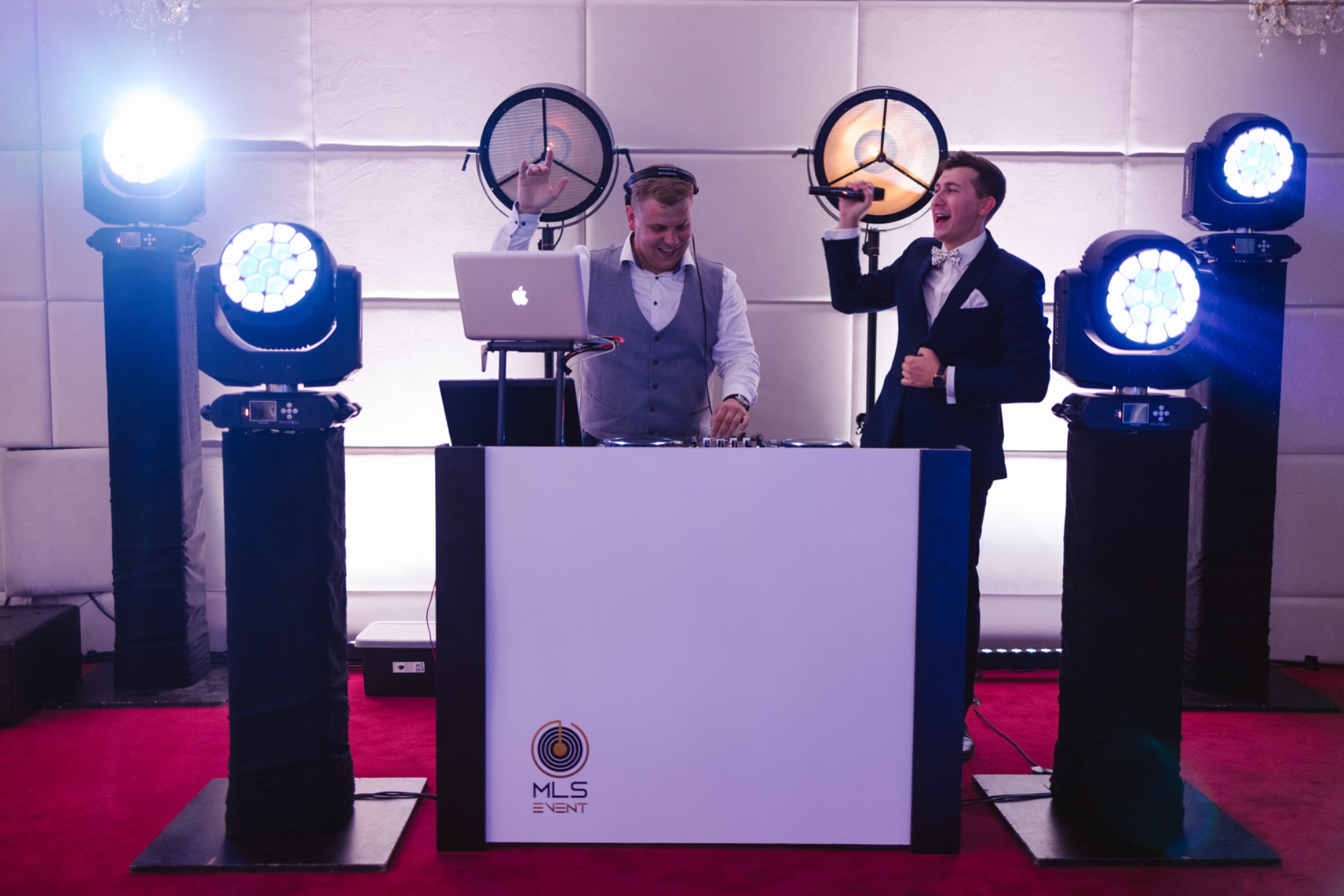 Uśmiechnięty DJ w akcji, obok wokalista w garniturze z mikrofonem. Scena oświetlona reflektorami, nowoczesny sprzęt DJ-ski, dekoracyjne tło.