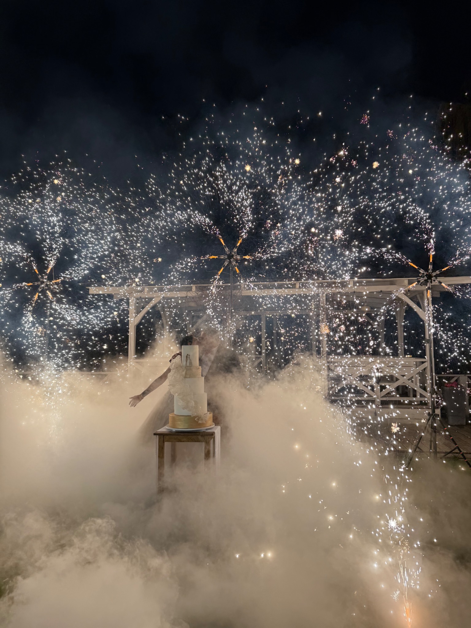 Tort weselny otoczony fontannami iskier i dymem, nocne niebo, drewniana altana w tle. Uroczysta oprawa wizualna, efekt specjalny.