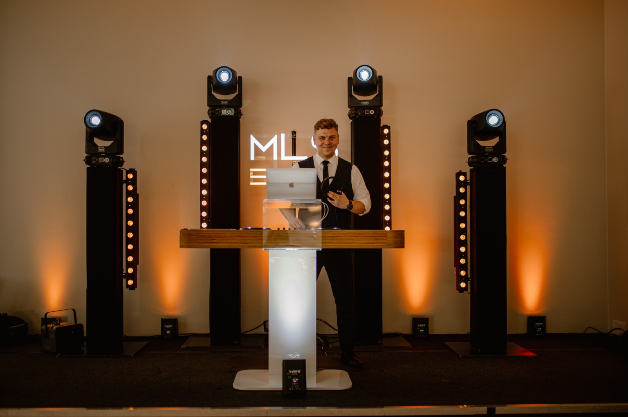 Uśmiechnięty DJ w kamizelce i krawacie, za konsoletą z laptopem, otoczony profesjonalnym oświetleniem scenicznym i neonowym logo. Scena gotowa na imprezę.