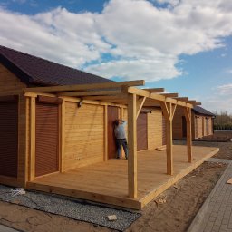 Firma Usługowo-Budowlana Wojciech Piatek - Montaż drewnianej altany z tarasem przy domku letniskowym. Widoczny pracownik podczas instalacji drzwi. W tle błękitne niebo z chmurami i fragment ścieżki.