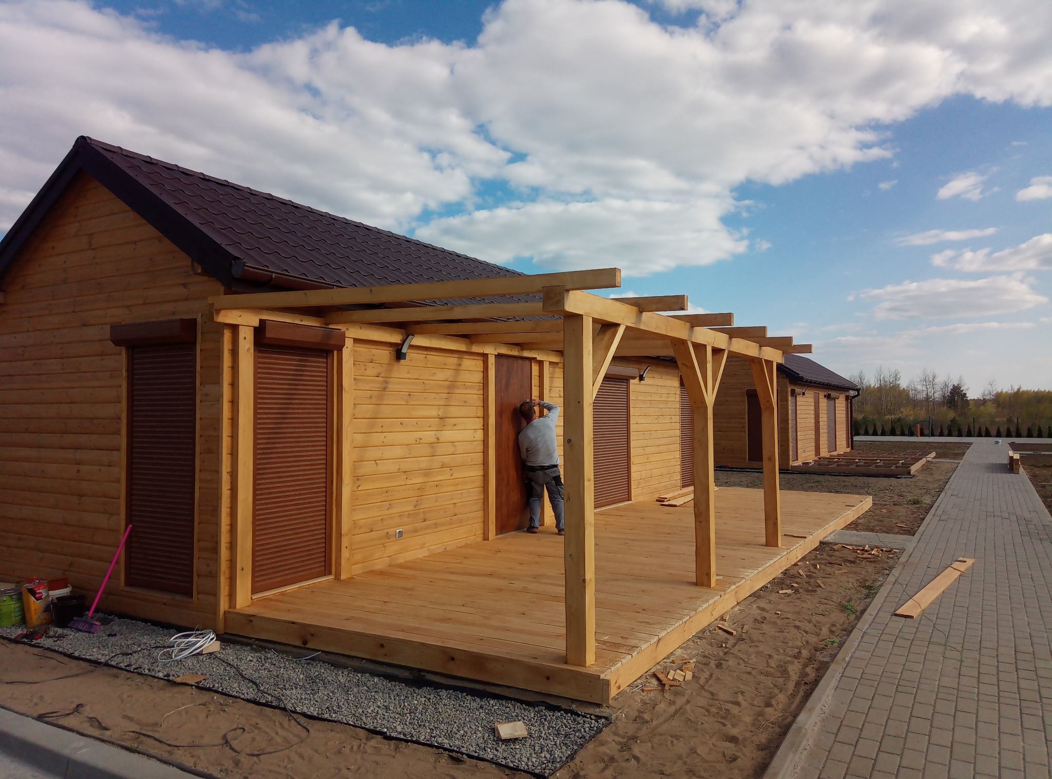 Montaż drewnianej altany z tarasem przy domku letniskowym. Widoczny pracownik podczas instalacji drzwi. W tle błękitne niebo z chmurami i fragment ścieżki.
