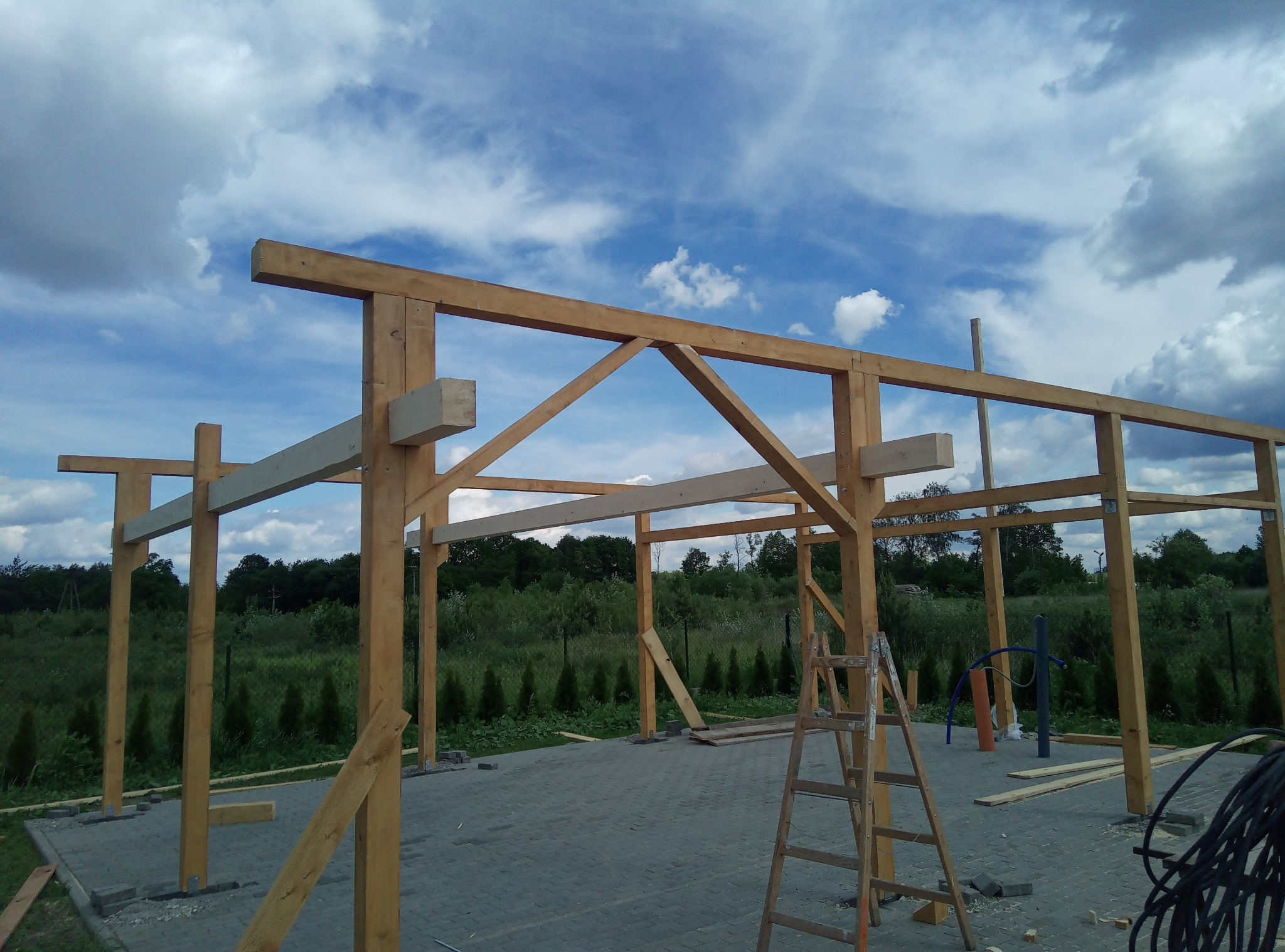 Wznoszona drewniana konstrukcja altany na kostce brukowej, z widoczną drabiną i zielonym tłem roślinności pod błękitnym niebem.