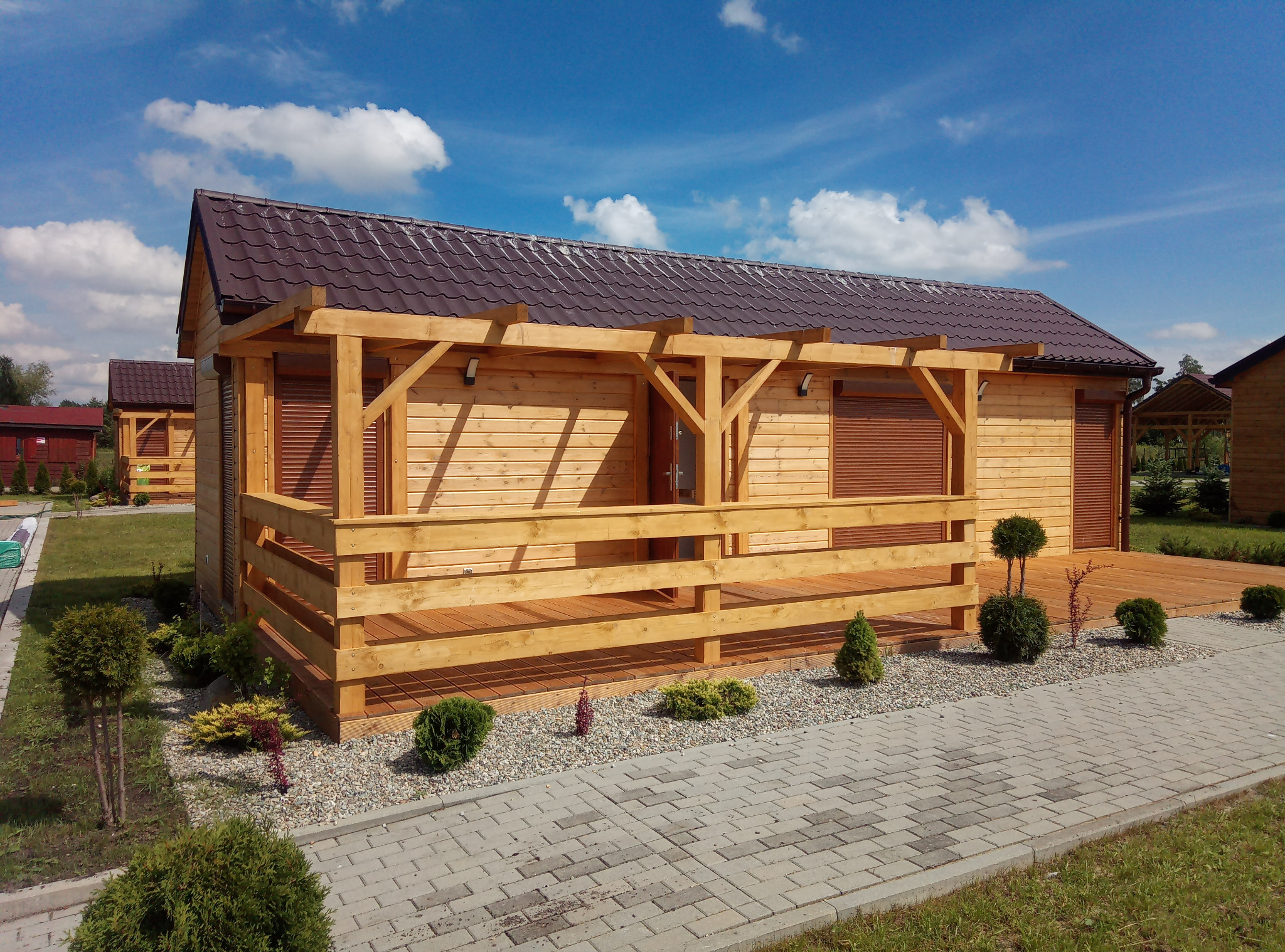 Drewniana altana z brązowym dachem i tarasem, otoczona zielenią i kostką brukową. Widoczne detale wykończenia i oświetlenie zewnętrzne.