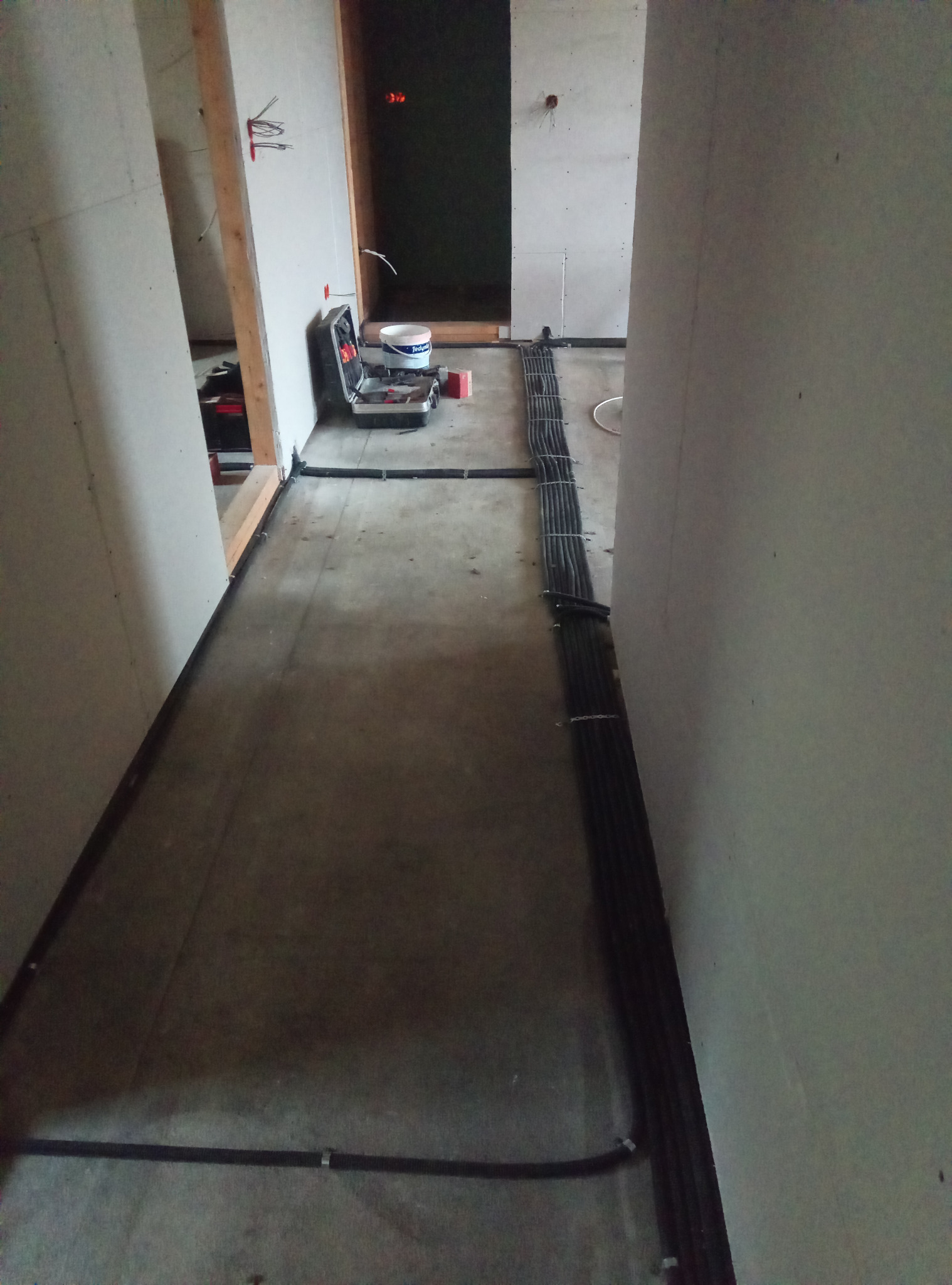 Kable elektryczne w peszlach ułożone na podłodze w korytarzu w trakcie prac wykończeniowych. Na ścianach płyty gipsowo-kartonowe, widoczna skrzynka narzędziowa.