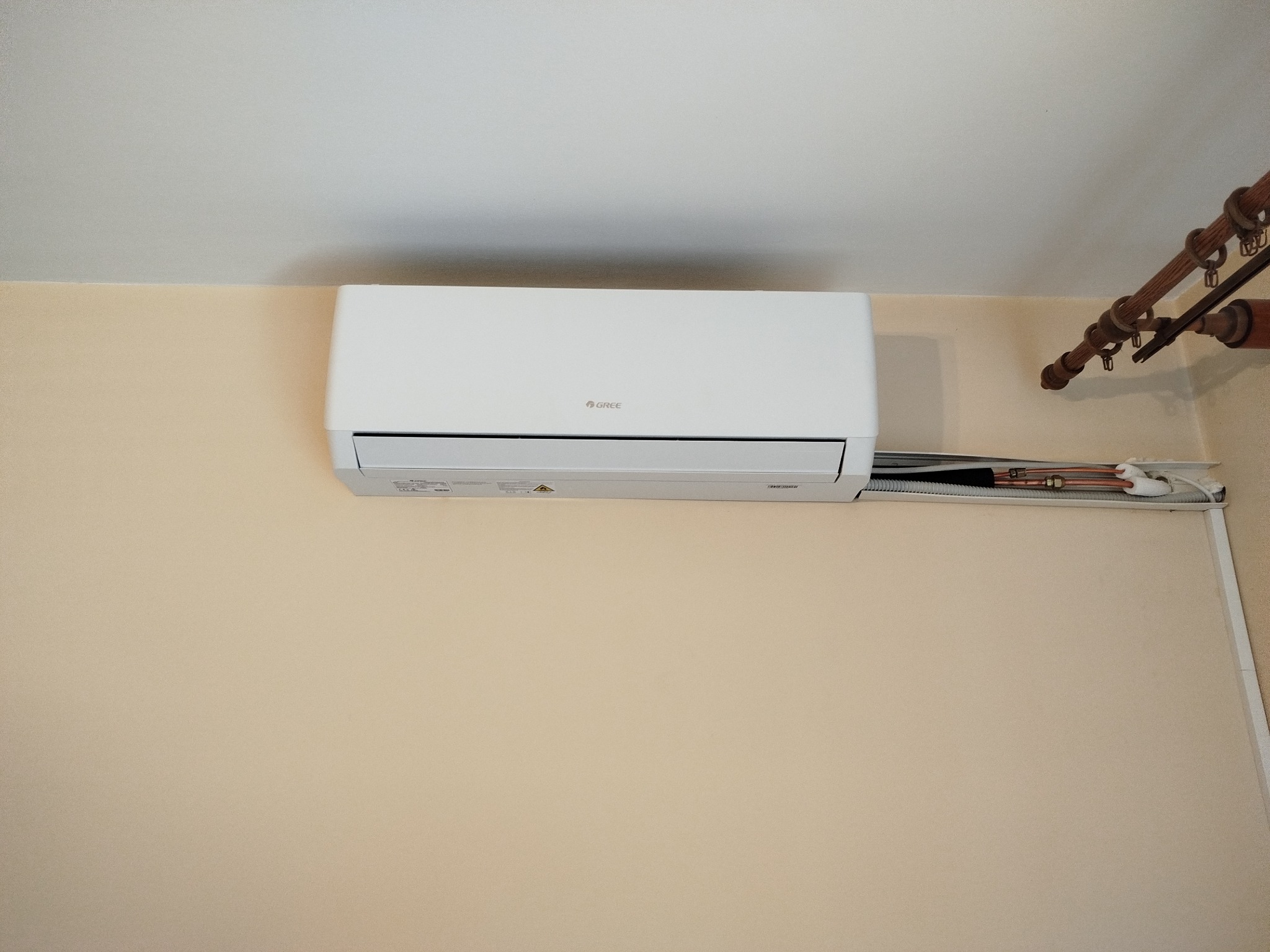 Biały klimatyzator marki Gree zamontowany na beżowej ścianie, widoczne rurki instalacyjne. U góry fragment karnisza. Ujęcie z dołu, centralne.