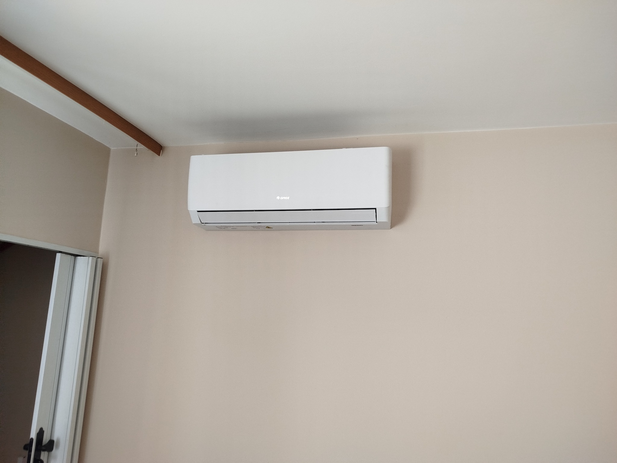 Biały klimatyzator marki Gree zamontowany na beżowej ścianie w pomieszczeniu. Widoczny fragment sufitu z listwą dekoracyjną i fragment drzwi.