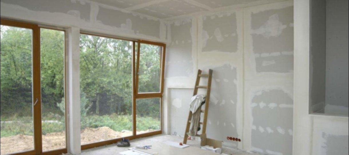 Pokój w trakcie szpachlowania ścian i sufitu, widoczna drabina oparta o ścianę, duże okna z widokiem na zieleń. Etap prac wykończeniowych, przygotowanie do malowania.