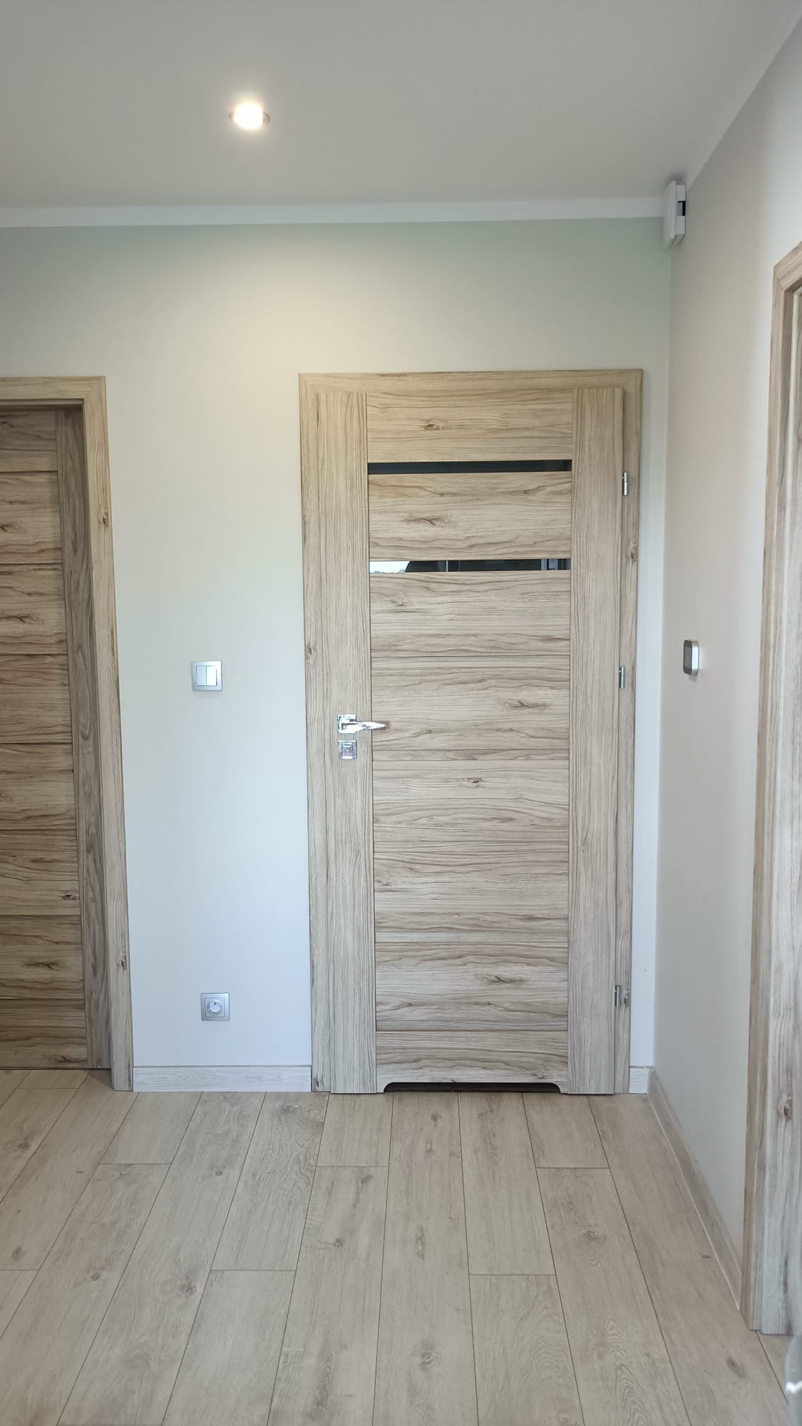 Jasne, drewnopodobne drzwi wewnętrzne z poziomymi panelami i przeszkleniem, zamontowane w świeżo wyremontowanym korytarzu z panelami podłogowymi.