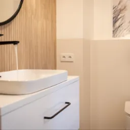 Nowoczesna łazienka: biała umywalka nablatowa z czarną armaturą, okrągłe lustro, beżowe płytki imitujące drewno i podwieszana toaleta.