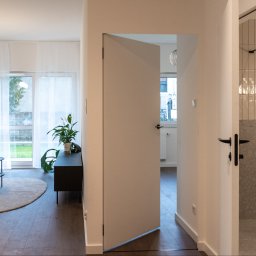 Ready Living REAPOLIS SP. Z O.O. DEV 21 SK - Jasne wnętrze mieszkania z otwartymi drzwiami, ukazującymi wykończoną łazienkę z mozaiką. Widoczny salon z sofą i oknem na ogród. Nowoczesny design.