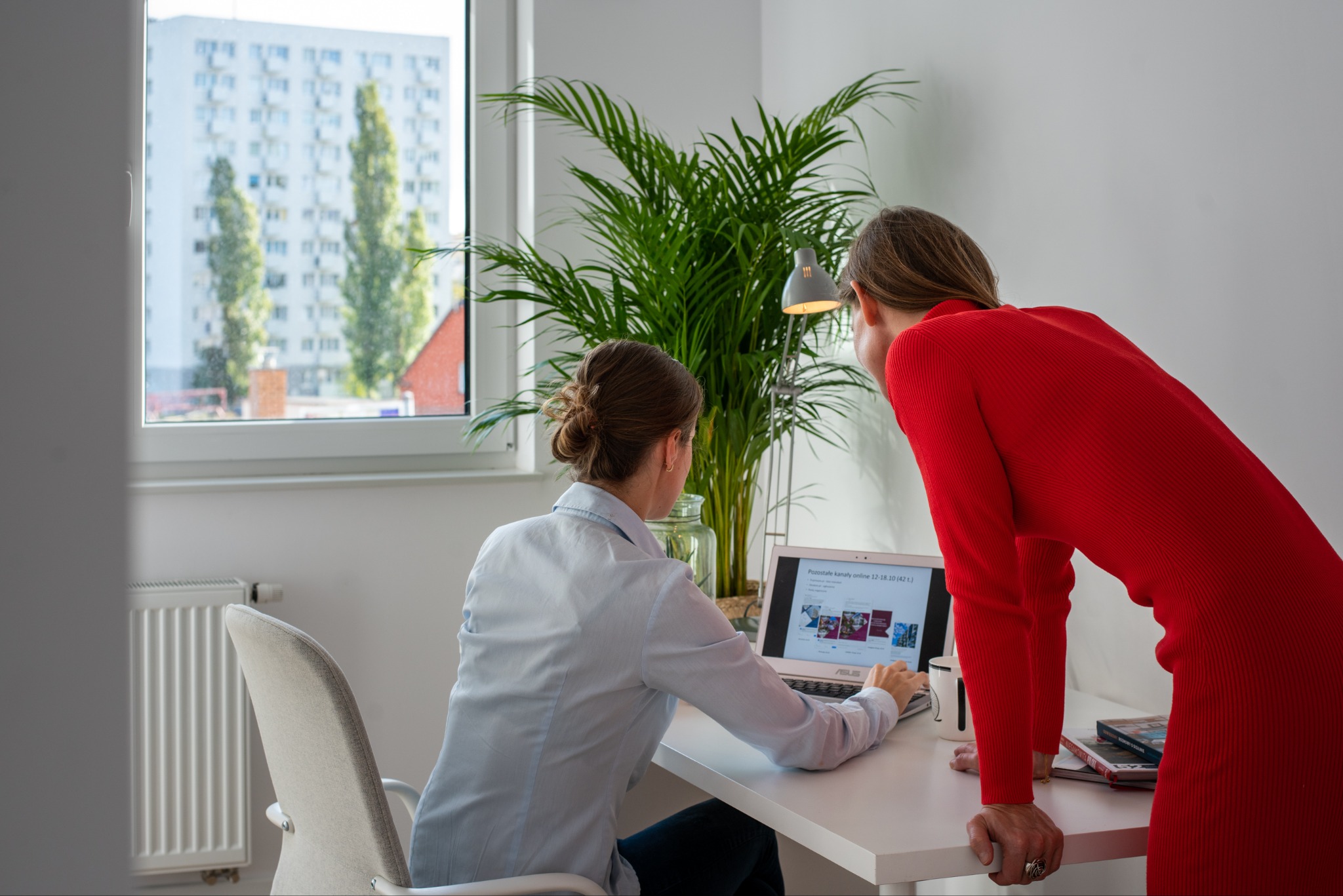 Dwie kobiety przeglądają laptopa w jasnym wnętrzu z widokiem na blok mieszkalny. Na biurku lampka, roślina doniczkowa i książki. Jedna osoba w czerwonej sukience.