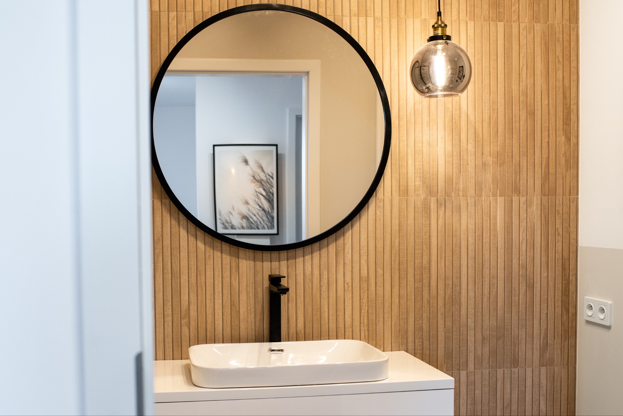 Nowoczesna łazienka z okrągłym lustrem i drewnopodobnymi płytkami. Umywalka nablatowa z czarną armaturą. Lampa wisząca ze szklanym kloszem.