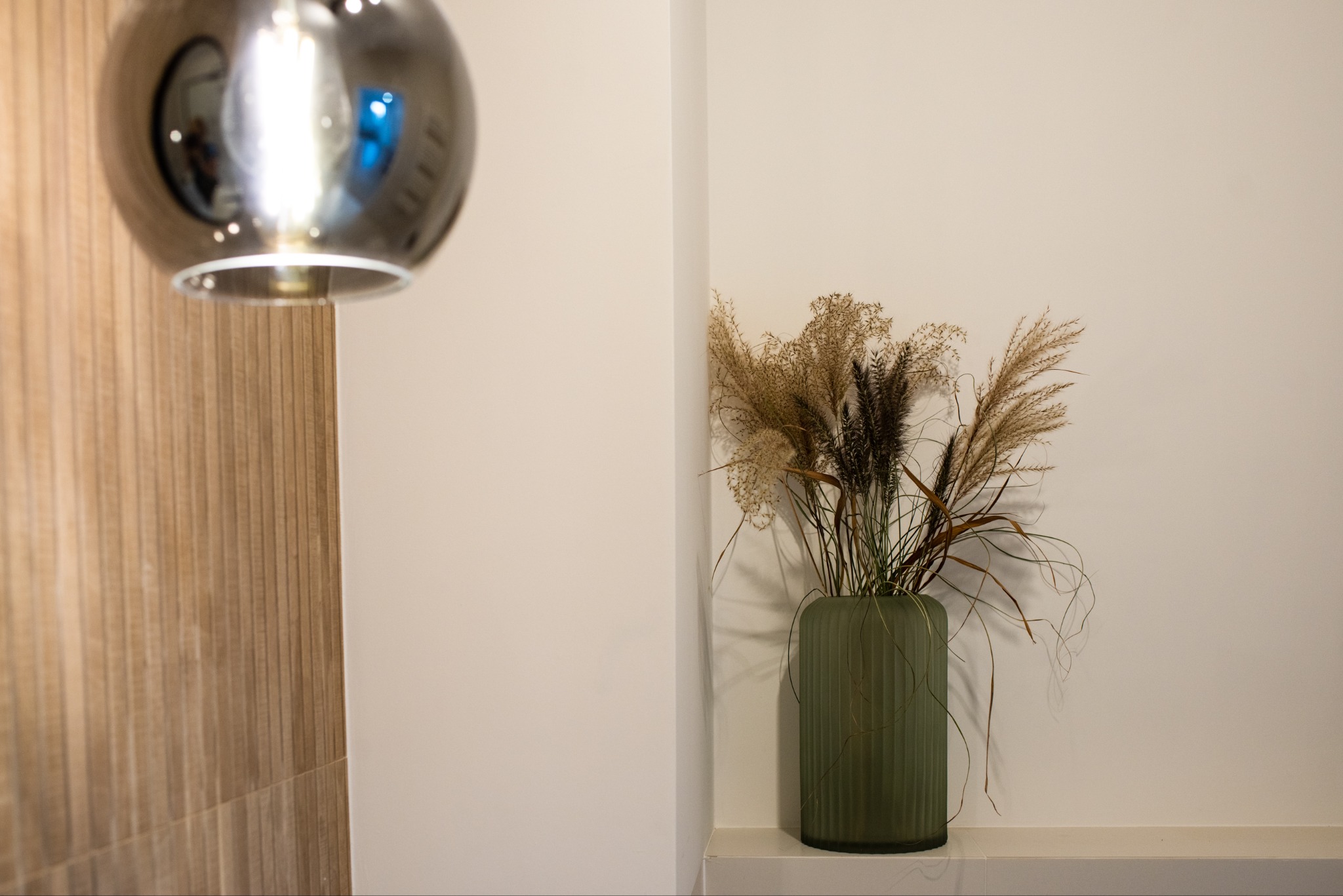 Dekoracyjny wazon z suszem na tle kremowej ściany i drewnopodobnych paneli. Nad nim zwisa okrągła, metalowa lampa, odbijająca otoczenie.