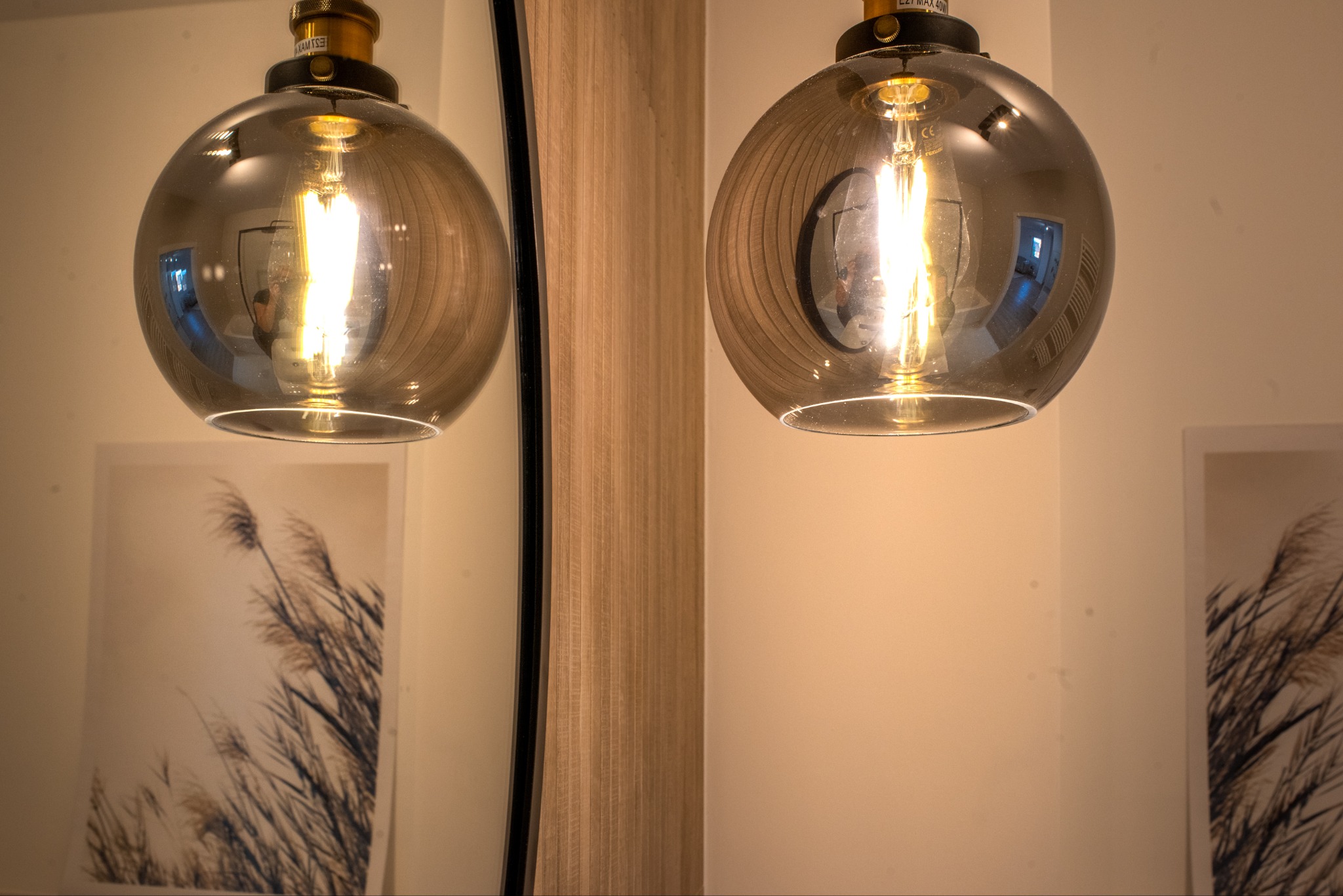 Dwie szklane lampy wiszące w kształcie kuli, z żarnikowymi żarówkami, oświetlające beżową ścianę z obrazem trzcin i lustrem. Ciepłe, nastrojowe światło.