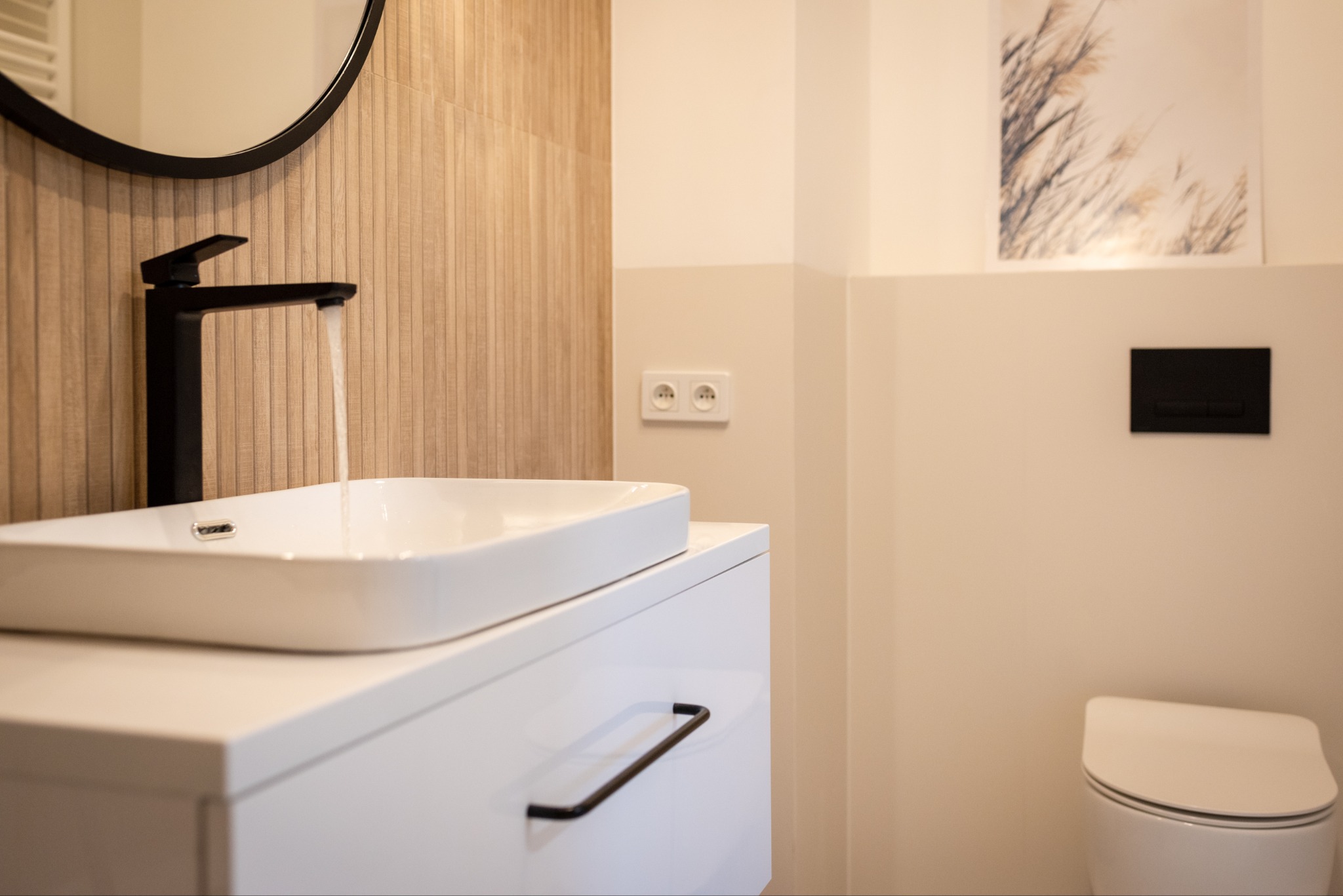 Nowoczesna łazienka: biała umywalka nablatowa z czarną armaturą, okrągłe lustro, beżowe płytki imitujące drewno i podwieszana toaleta.