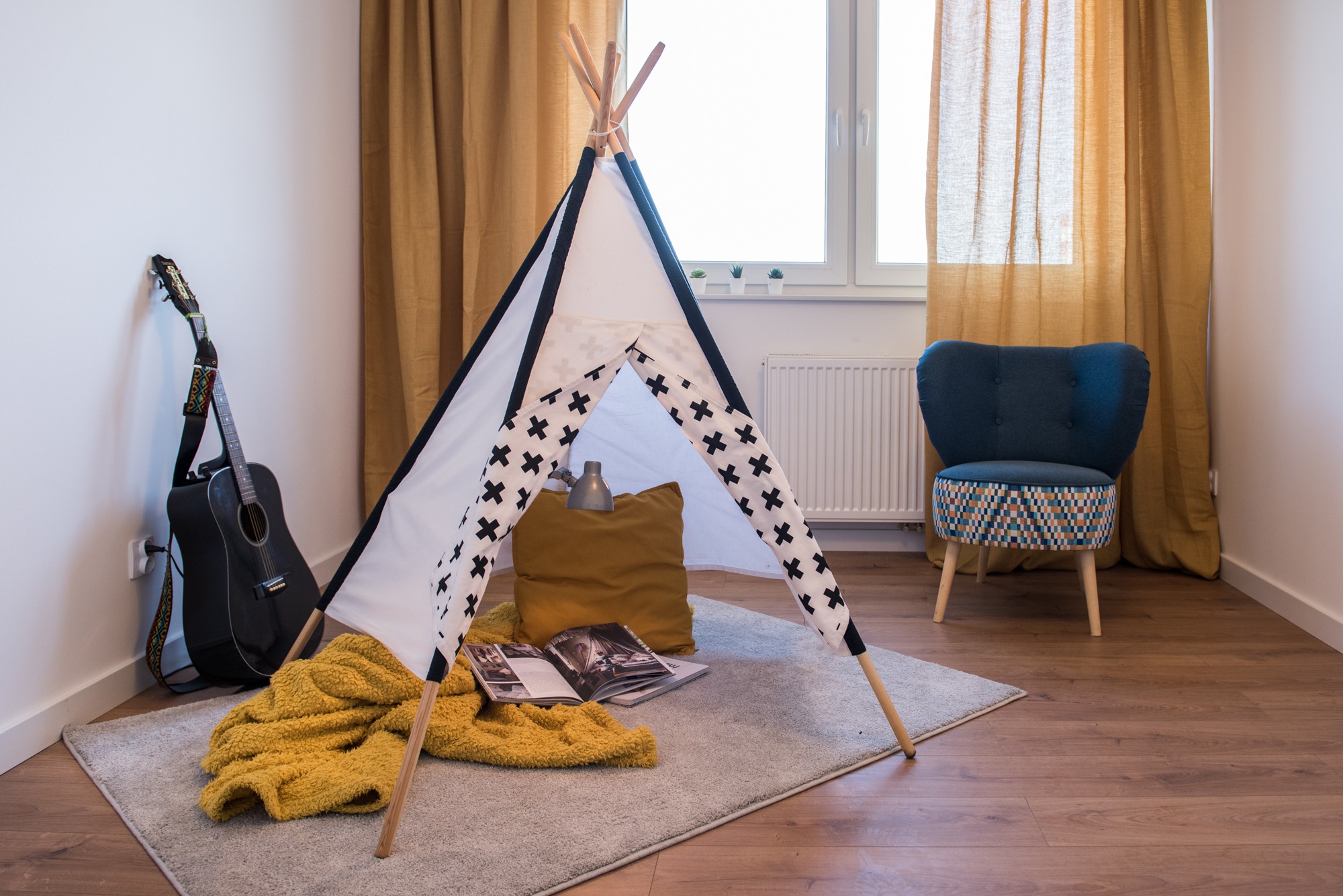 Pokój dziecięcy z tipi w stylu boho: biało-czarny namiot z krzyżami, gitara oparta o ścianę, pluszowy koc i niebieski fotel na drewnianej podłodze.