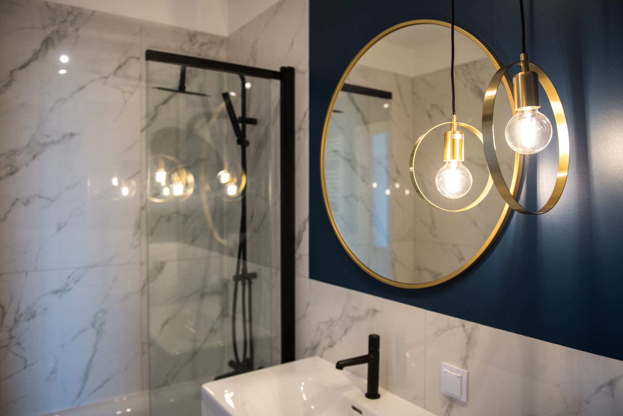 Nowoczesna łazienka: kabina prysznicowa z czarnymi ramami, okrągłe lustro w złotej ramie i designerskie lampy wiszące. Marmurowe płytki i granatowa ściana.