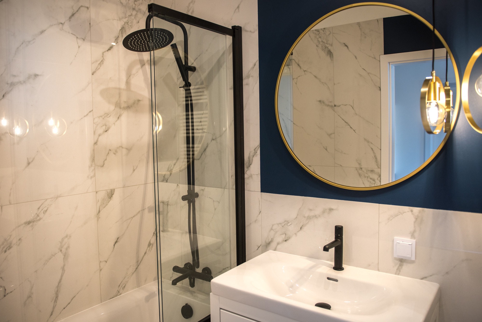 Nowoczesna łazienka z kabiną prysznicową typu walk-in z czarną armaturą, okrągłym lustrem w złotej ramie i marmurowymi płytkami. Minimalistyczny design.