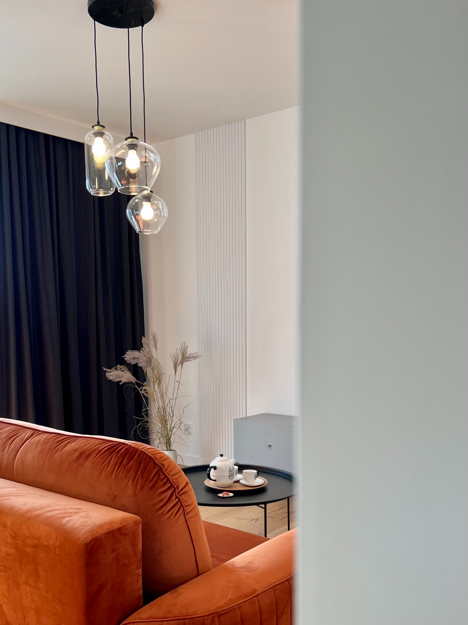 Stylowe wnętrze z pomarańczową sofą, okrągłym stolikiem z serwisem do herbaty, wiszącymi lampami i czarnymi zasłonami. Dekoracyjna ściana z pionowymi panelami.