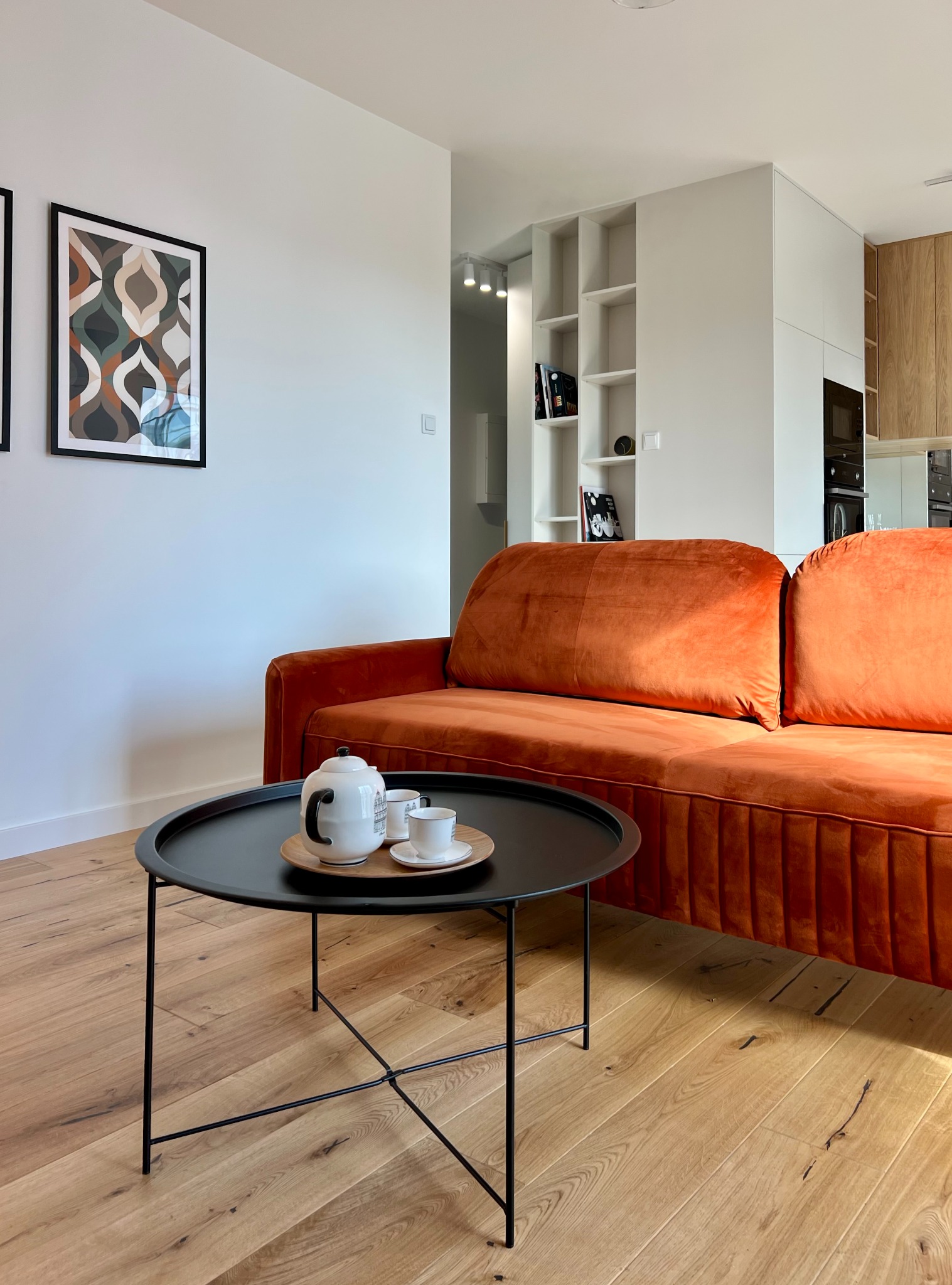 Stylowy salon z pomarańczową sofą, czarnym stolikiem kawowym i drewnianą podłogą. Na ścianie wiszą grafiki w czarnych ramkach. W tle widoczna zabudowa kuchenna.