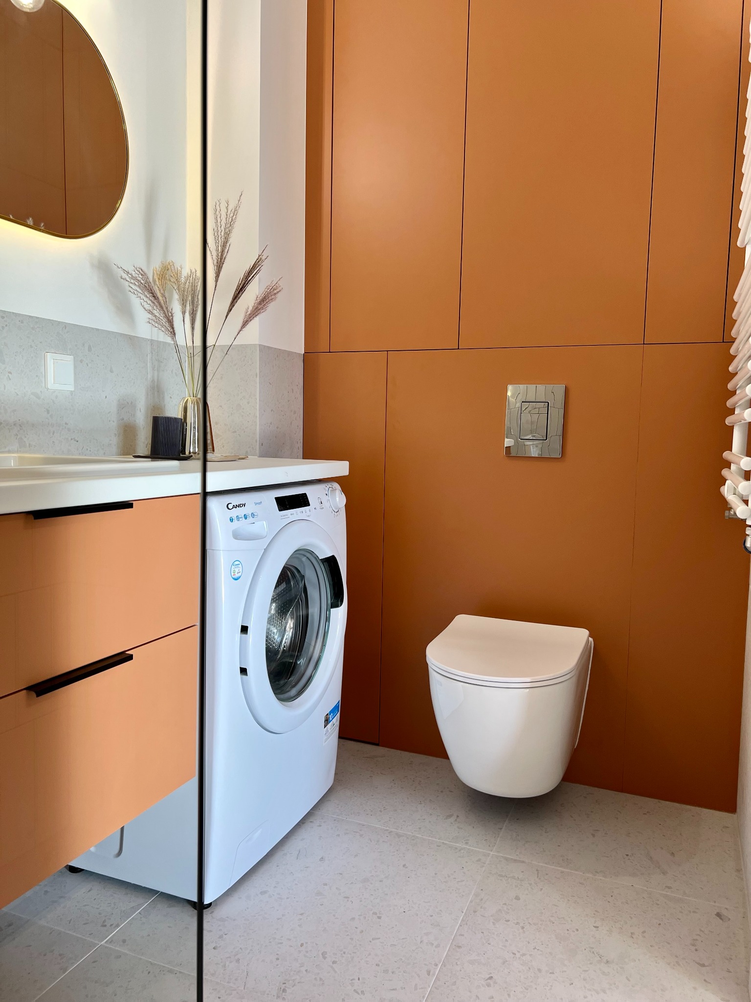 Pomarańczowa zabudowa w łazience z umywalką, pralką i podwieszaną toaletą. Uwagę zwracają minimalistyczne, czarne uchwyty i szare płytki podłogowe.