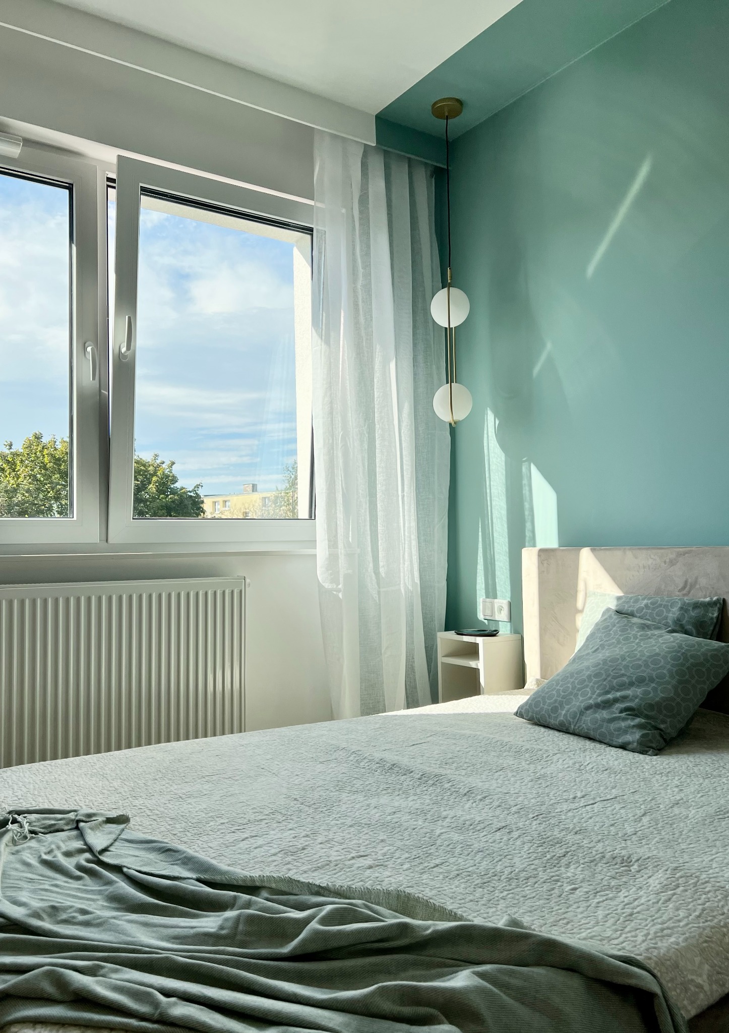 Jasne wnętrze sypialni z turkusową ścianą, białym oknem z widokiem na zieleń i łóżkiem z poduszkami w odcieniach szarości oraz lampą wiszącą.