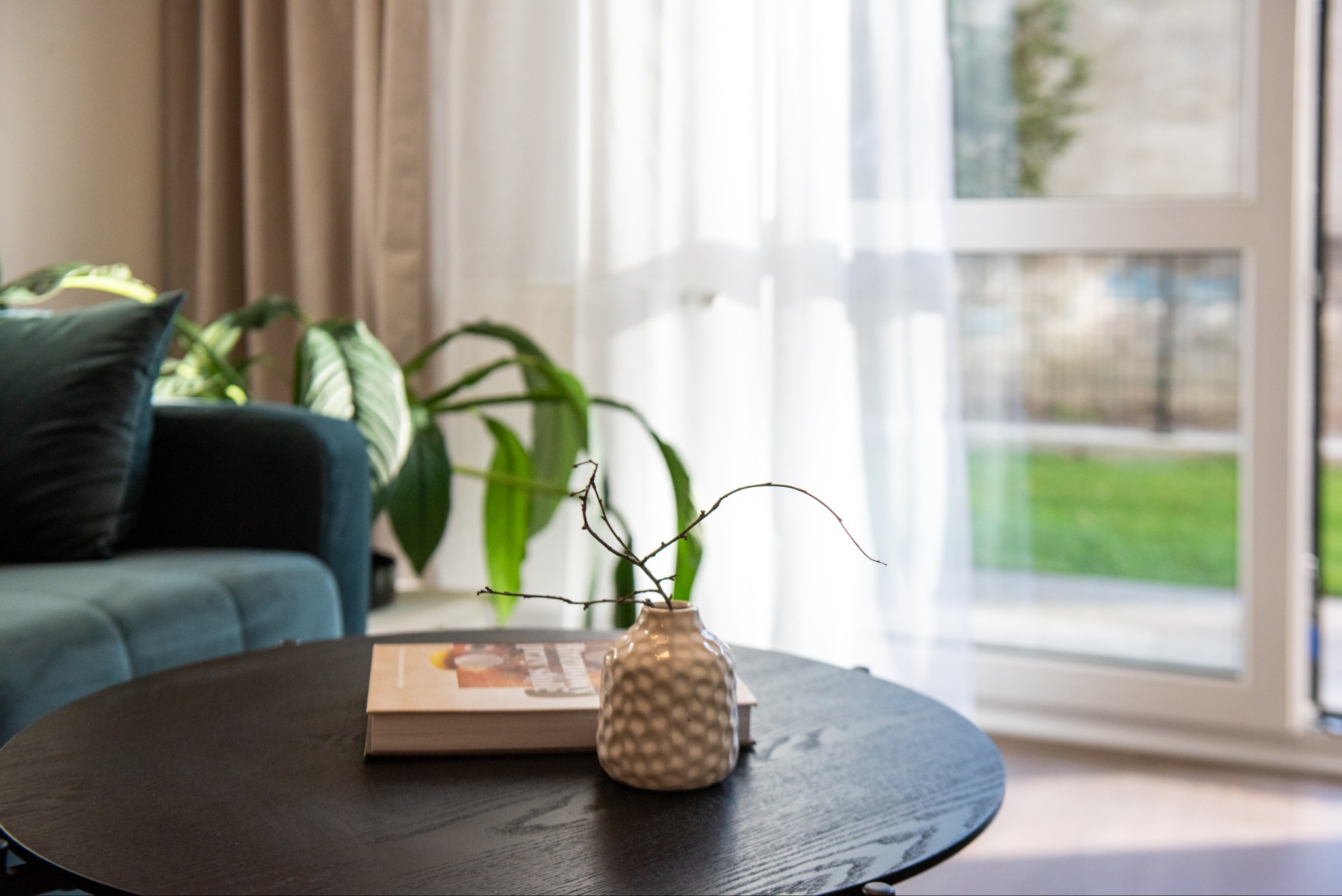 Stylowy salon z turkusową sofą i okrągłym stolikiem. Na blacie wazon z gałązkami i książka. W tle okno z białymi firanami i widok na zieleń.