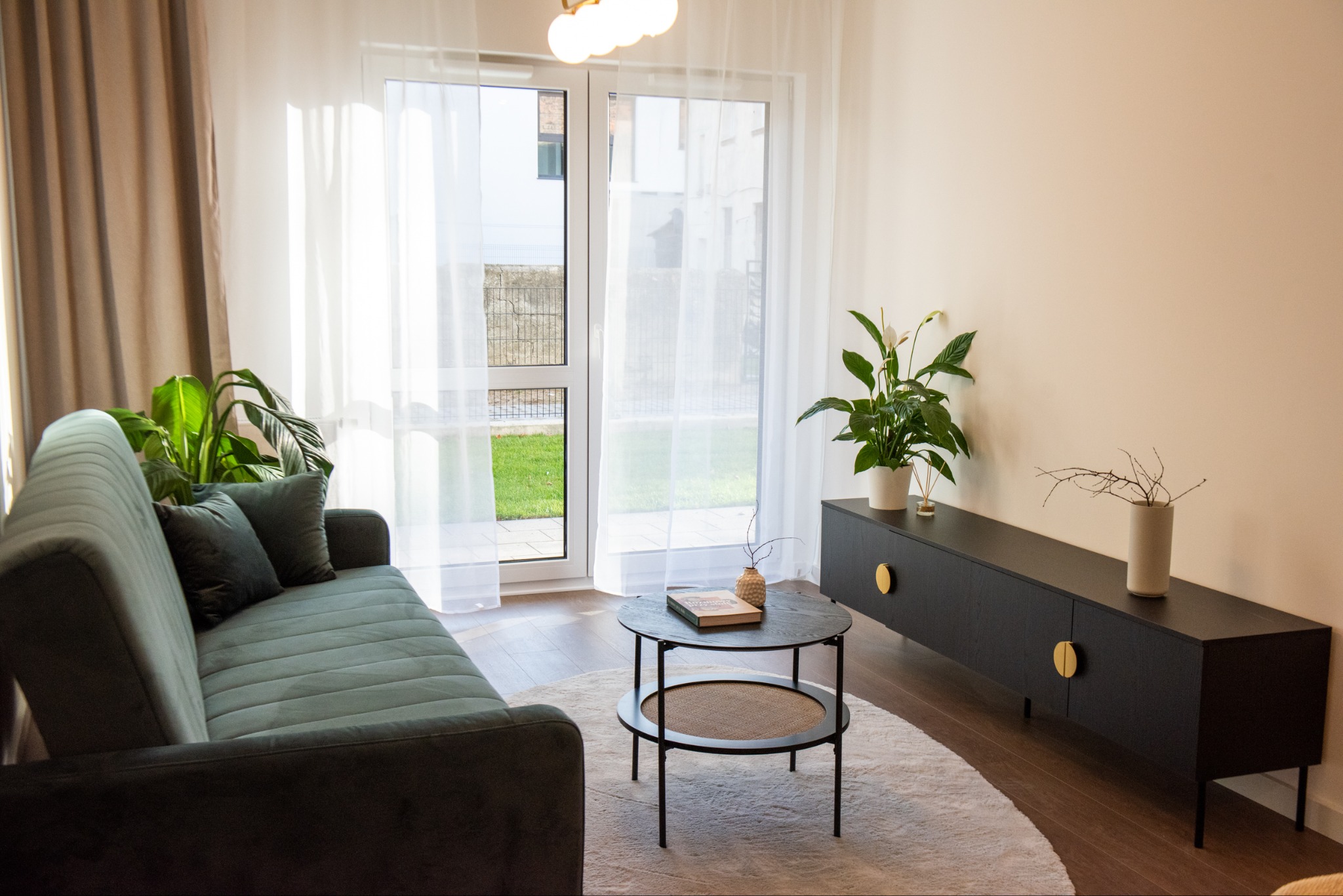 Jasne wnętrze salonu z zieloną sofą, okrągłym stolikiem i czarną komodą z roślinami. Duże okno balkonowe z białymi zasłonami wpuszcza naturalne światło.