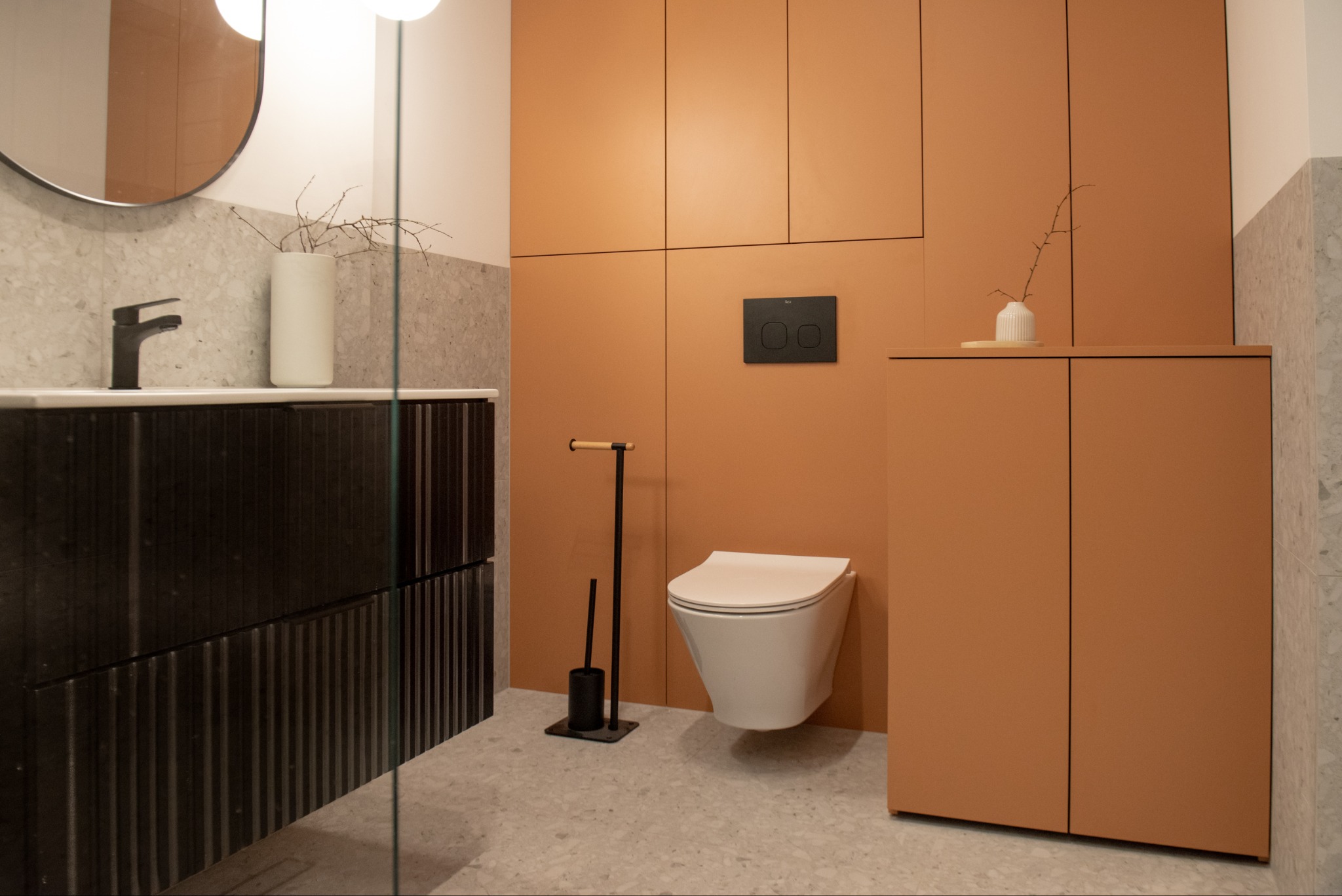 Nowoczesna łazienka z wiszącą toaletą, umywalką z ciemnego drewna i terakotowymi szafkami. Minimalistyczny design z akcentami dekoracyjnymi.