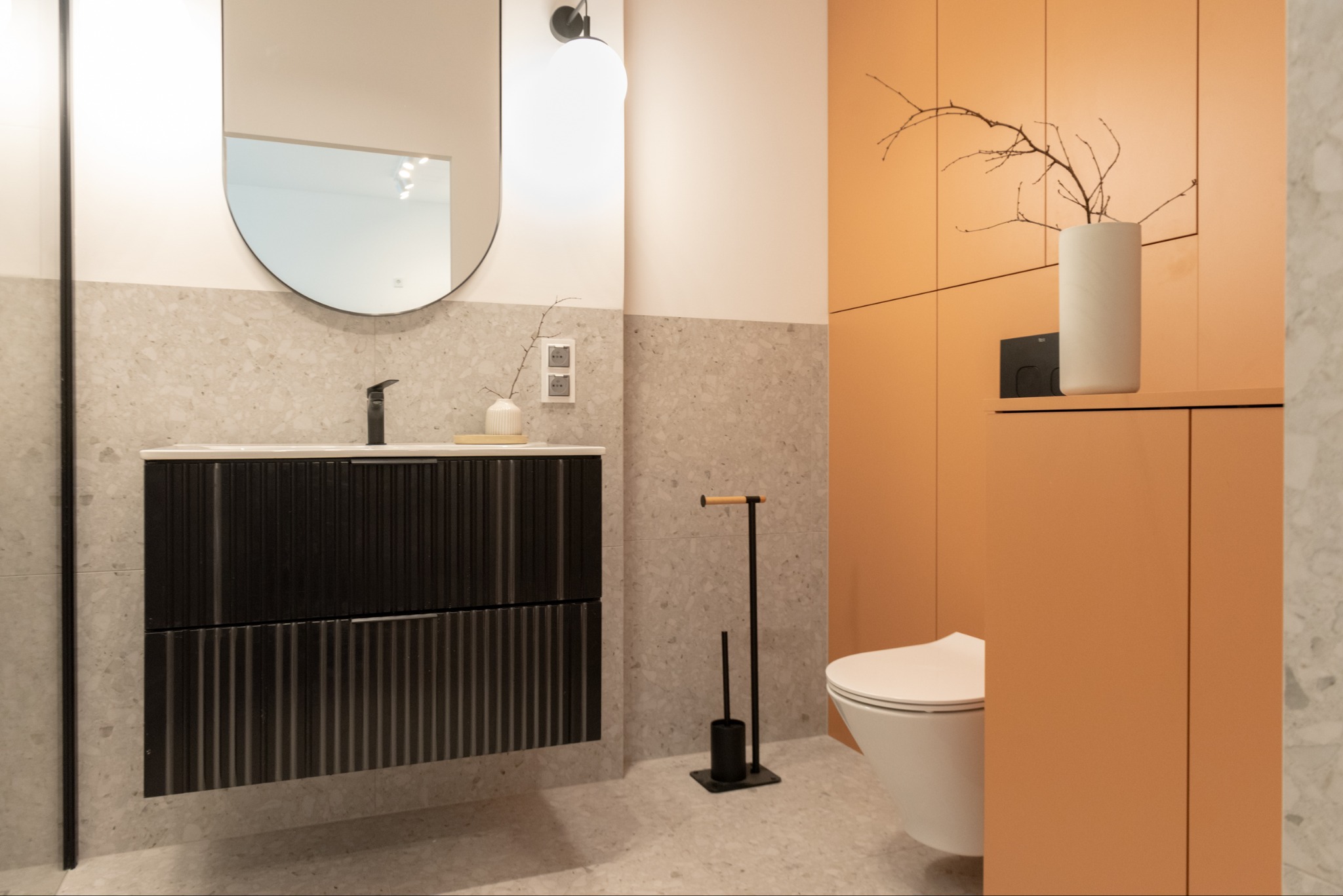 Nowoczesna łazienka w beżach i pomarańczach: szafka z umywalką, lustro z lampą, toaleta w zabudowie, dekoracje z gałązek. Minimalistyczny design.