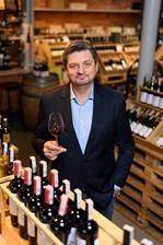 Mężczyzna w garniturze trzyma kieliszek czerwonego wina na tle półek z butelkami w hurtowni alkoholi. Widoczny asortyment i osoba.