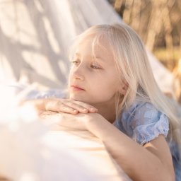 spoorna_m_photo Martyna Spórna - Portret dziewczynki o jasnych włosach, ubranej w błękitną sukienkę, leżącej na jasnej tkaninie na tle drzew w ciepłym świetle słonecznym. Delikatne ujęcie.