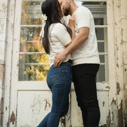 spoorna_m_photo Martyna Spórna - Zakochana para całuje się na tle białych, odrapanych drzwi z dekoracyjnym łukiem. Ona ma kokardę we włosach, oboje w białych koszulkach i sportowych butach.