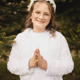 spoorna_m_photo Martyna Spórna - Portret dziewczynki w okularach, wianku i białej albie, z dłońmi złożonymi do modlitwy, na tle zielonego iglastego drzewa. Ujęcie z bliska, ciepłe światło.