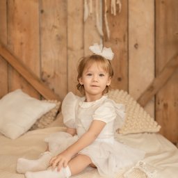 spoorna_m_photo Martyna Spórna - Portret dziewczynki w białej sukience, siedzącej na łóżku z drewnianym zagłówkiem i łapaczem snów w tle. Delikatne, ciepłe światło.