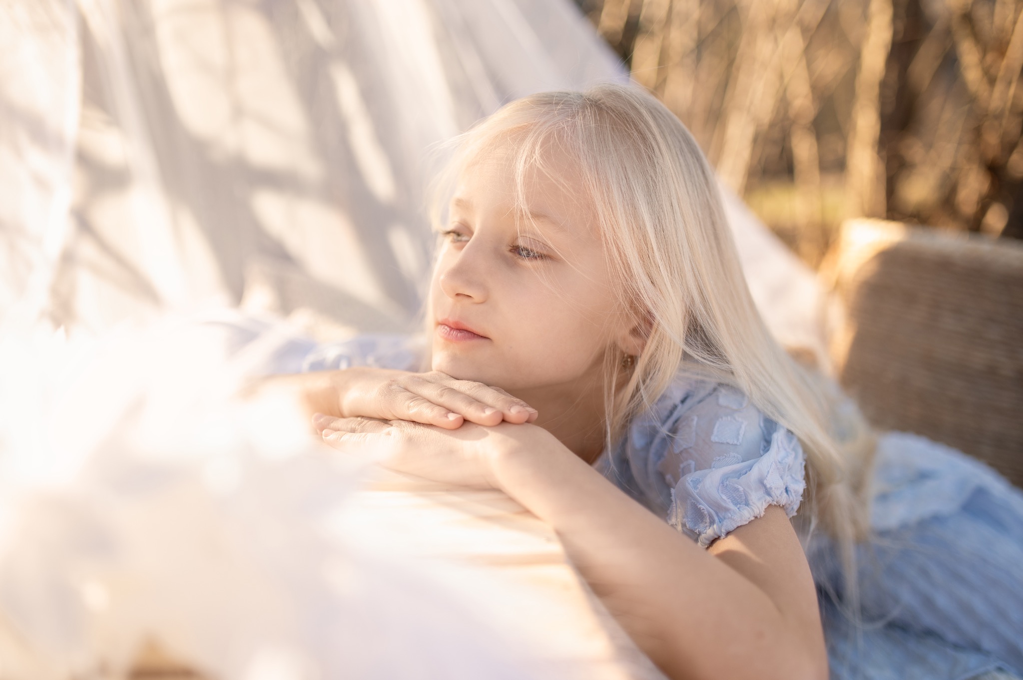 Portret dziewczynki o jasnych włosach, ubranej w błękitną sukienkę, leżącej na jasnej tkaninie na tle drzew w ciepłym świetle słonecznym. Delikatne ujęcie.
