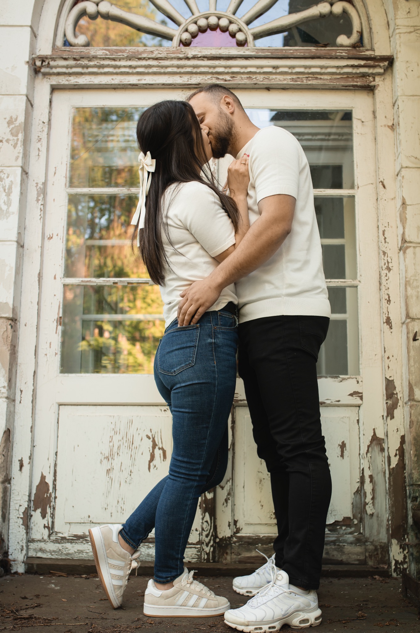 Zakochana para całuje się na tle białych, odrapanych drzwi z dekoracyjnym łukiem. Ona ma kokardę we włosach, oboje w białych koszulkach i sportowych butach.