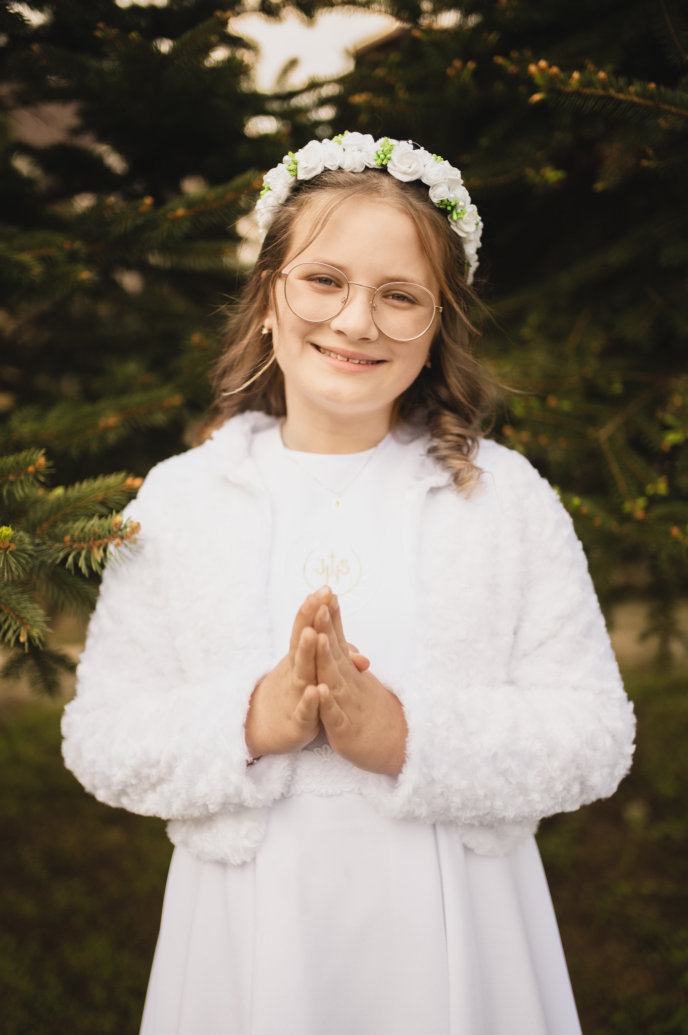 Portret dziewczynki w okularach, wianku i białej albie, z dłońmi złożonymi do modlitwy, na tle zielonego iglastego drzewa. Ujęcie z bliska, ciepłe światło.