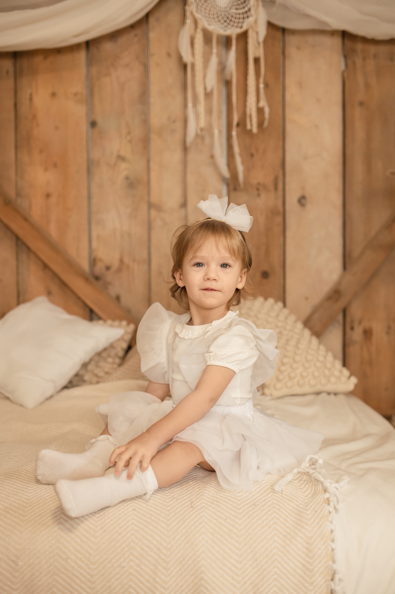 Portret dziewczynki w białej sukience, siedzącej na łóżku z drewnianym zagłówkiem i łapaczem snów w tle. Delikatne, ciepłe światło.