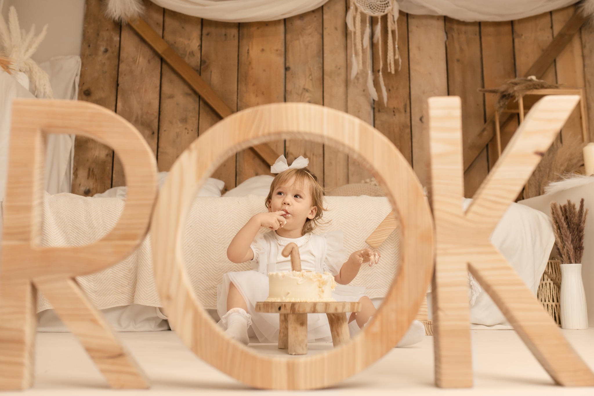 Roczne dziecko w białej sukience je tort z cyfrą 1, siedząc na małym stołku. W tle drewniane litery układają się w słowo 'ROK'. Scena w ciepłych, naturalnych barwach.