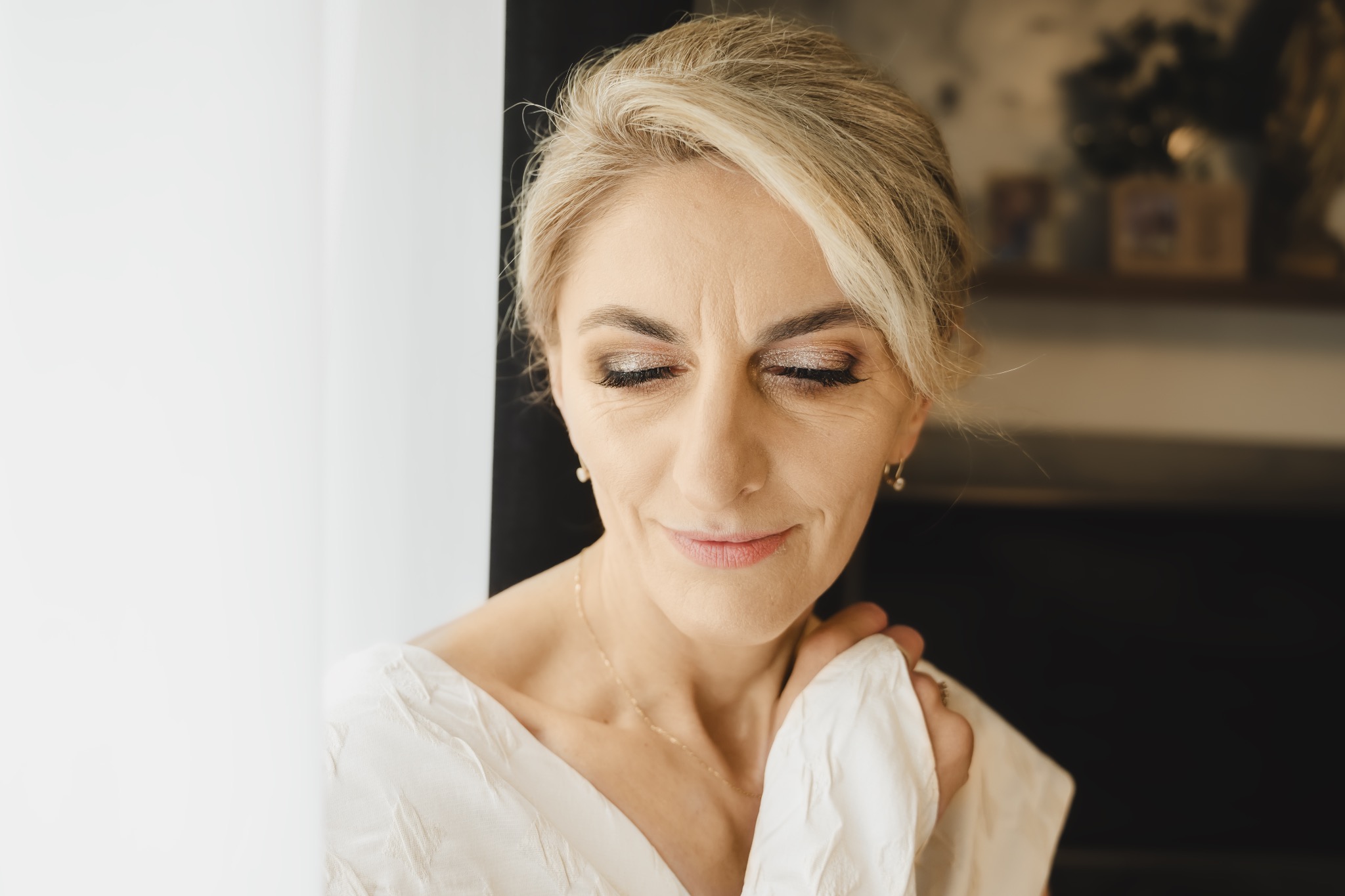 Portret kobiety o blond włosach z zamkniętymi oczami, delikatny makijaż i biała sukienka, uchwycona w miękkim świetle w pobliżu okna.