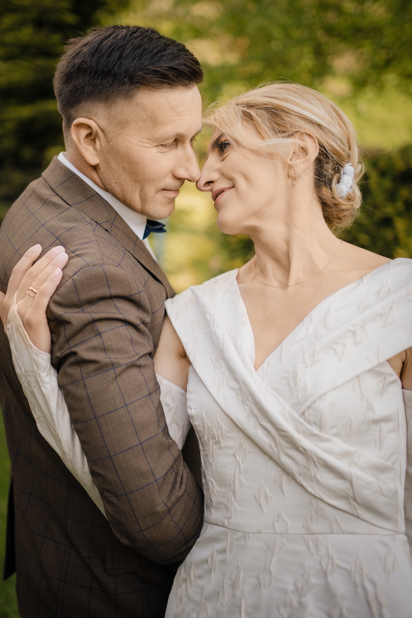 Uśmiechnięta para w zbliżeniu, dotykająca się nosami w plenerze. Kobieta w białej sukni, mężczyzna w brązowym garniturze w kratę i muszce.