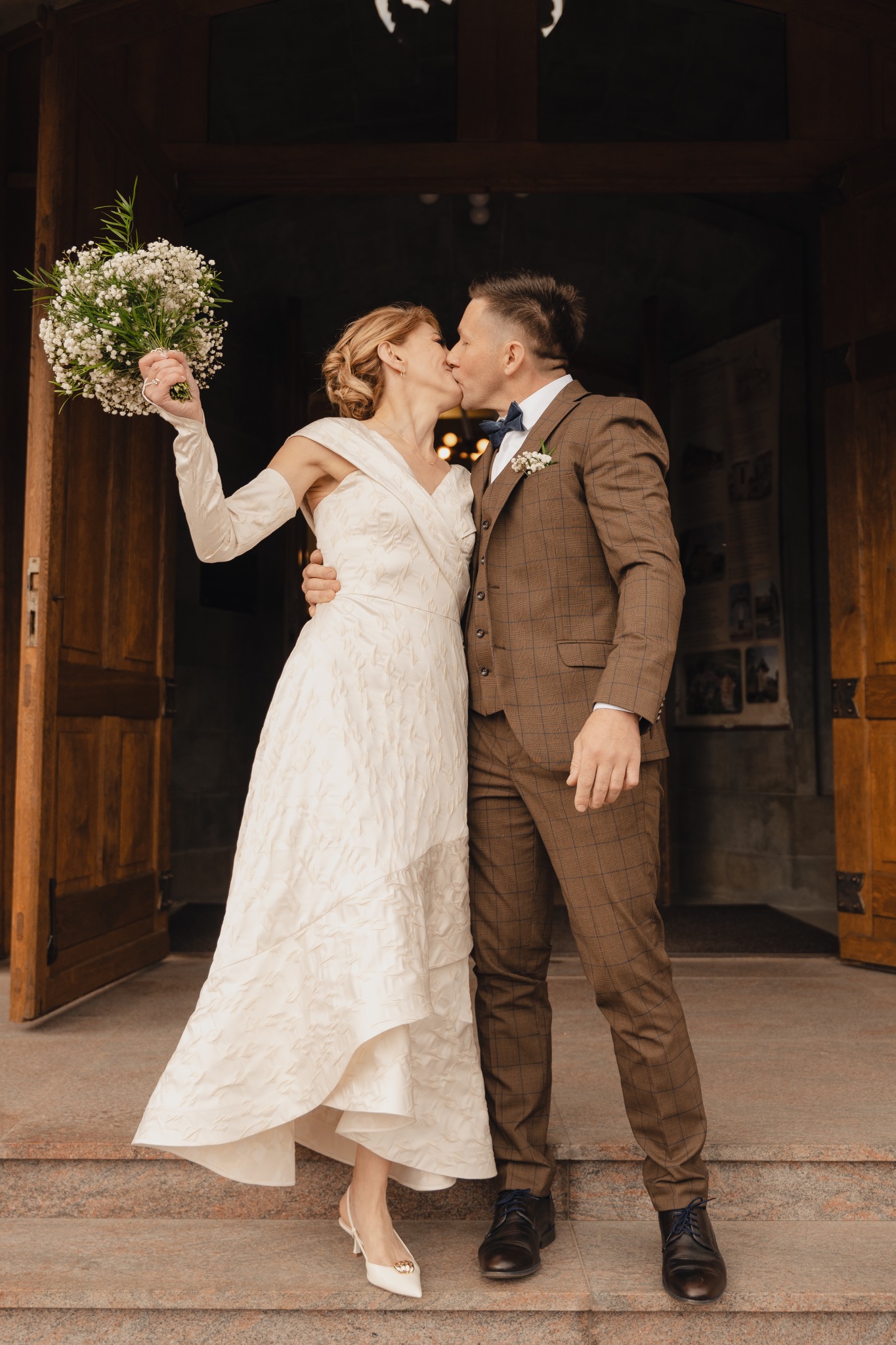 Nowożeńcy wymieniają pocałunek przed drewnianymi drzwiami; panna młoda w sukni ecru trzyma bukiet białych kwiatów, pan młody w brązowym garniturze w kratę.