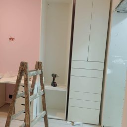 Artur Rzemiszke Usługi Remontowo-Wykończeniowe - Montaż białych szaf w pokoju z różową ścianą. Widoczna drabina, wiertarka na półce. Podłoga z paneli. Częściowo widoczna płyta karton-gips.